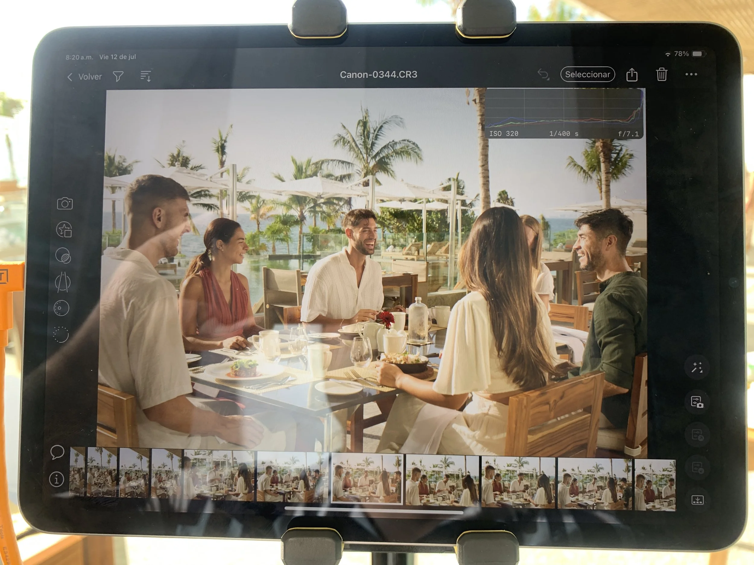 Group of friends laughing and talking at a table outdoors near a beach with palm trees.