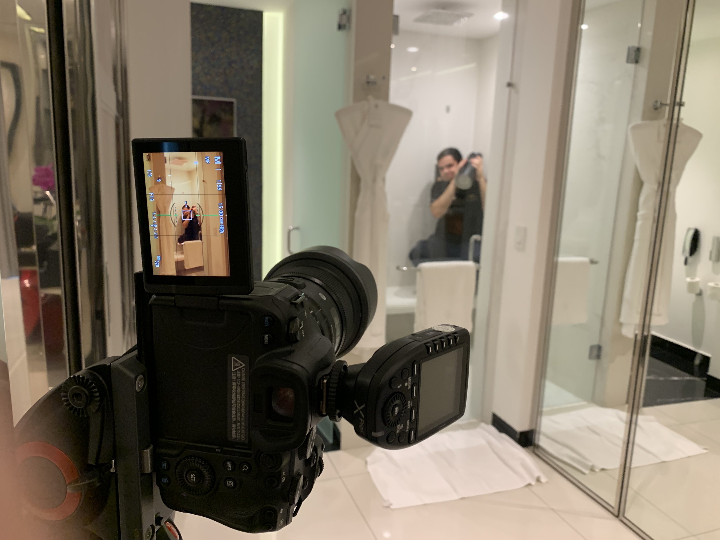 Camera capturing a person taking a photo of themselves in a mirror in a hotel bathroom with glass walls and towels on the floor.