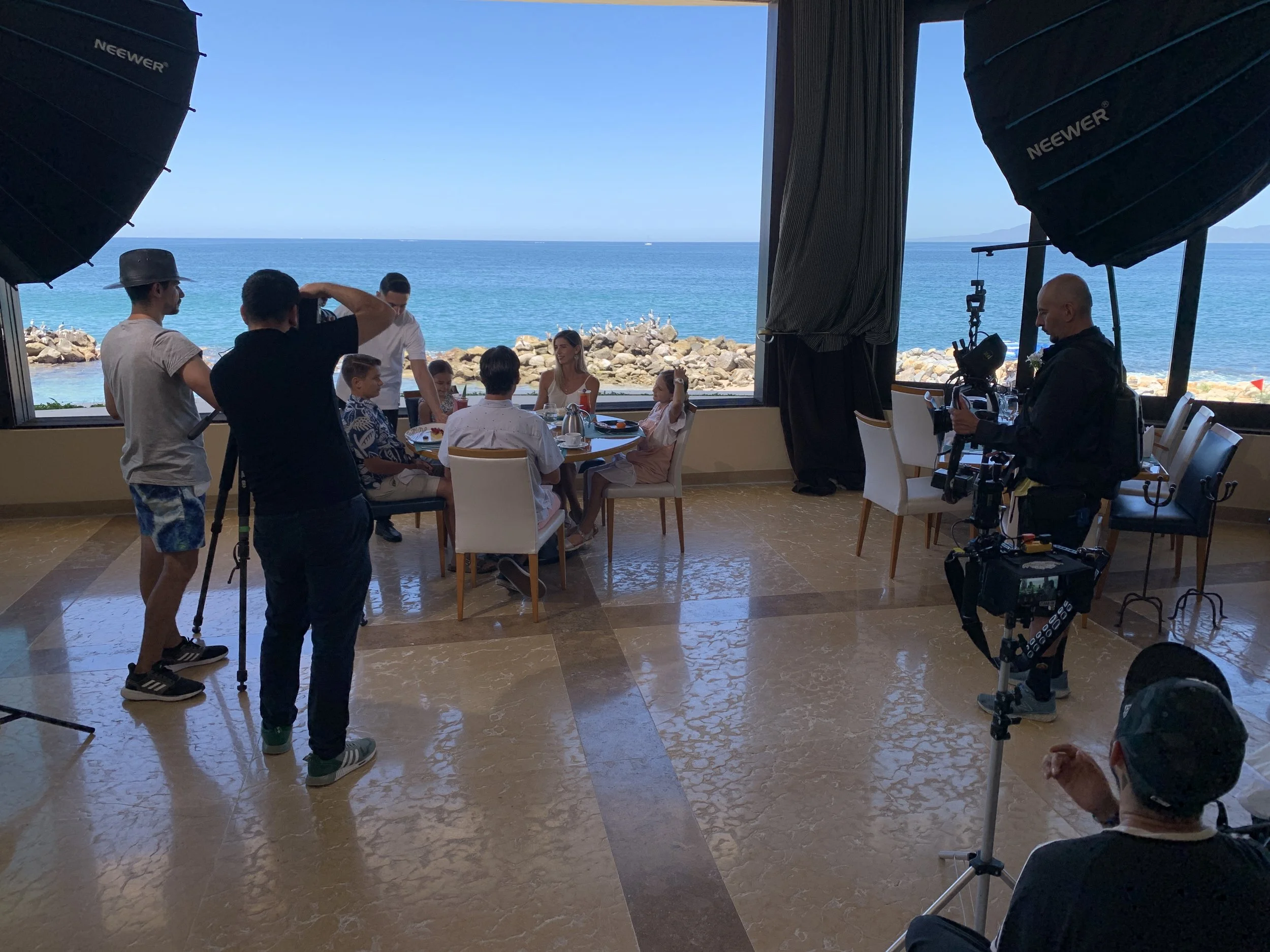 A photo captures a scene of a film or photo shoot on a seaside balcony with a view of the ocean and rocks. Several crew members are present, including a camera operator with equipment, and a photographer with a tripod. In the background, a group of seven people, including children and adults, are sitting at a round dining table set outdoors, engaging in a discussion or meal, with the ocean visible through large windows behind them.