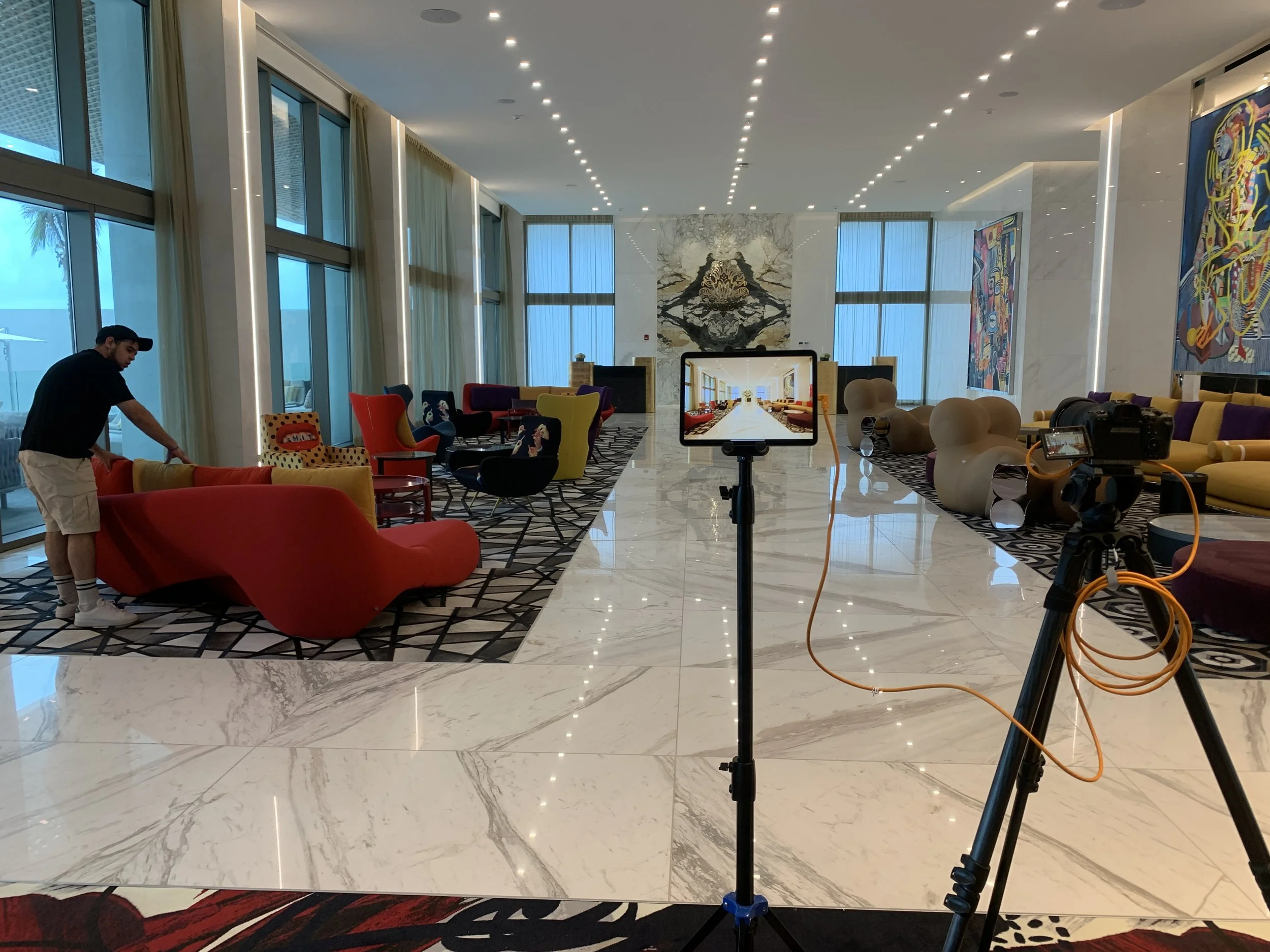 Colorful modern lobby with vibrant sofas and chairs, large windows with curtains, and artwork on the walls. A person is adjusting a red sofa, and filming equipment is set up in the foreground.