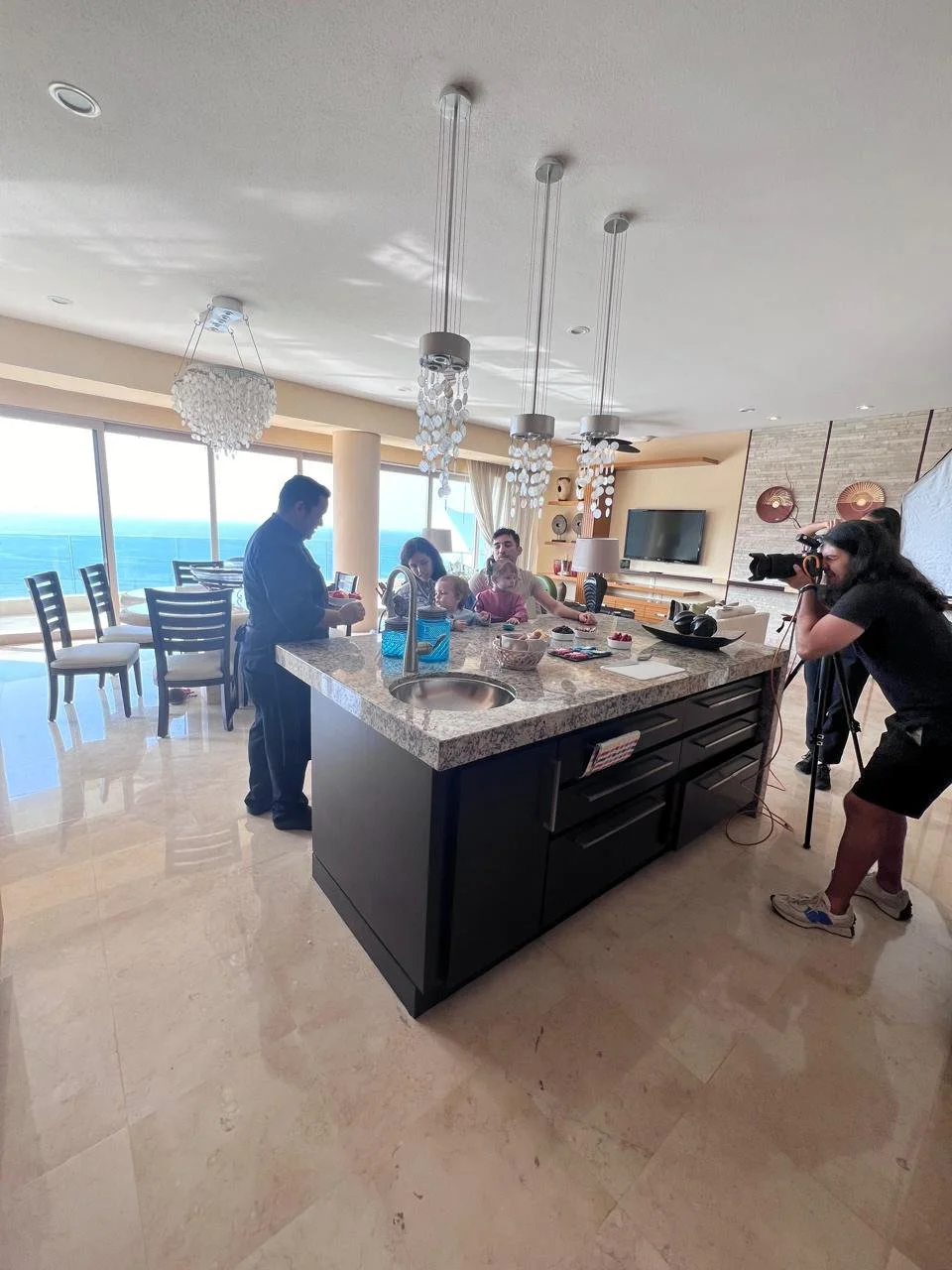 Family photoshoot being captured in a bright, modern kitchen with a large island, overlooking the ocean through large windows.
