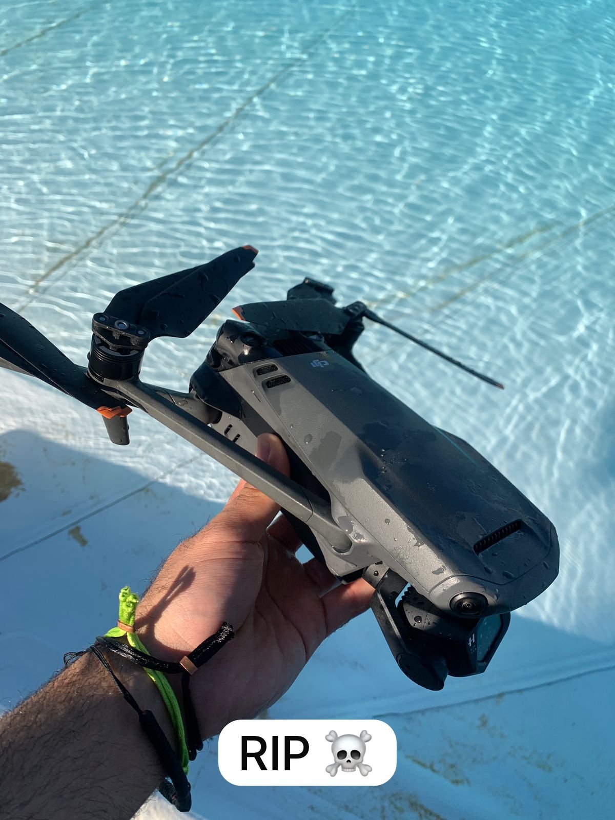 A person holding a damaged drone near a shallow, rippled blue water body, with a RIP skull emoji overlay.