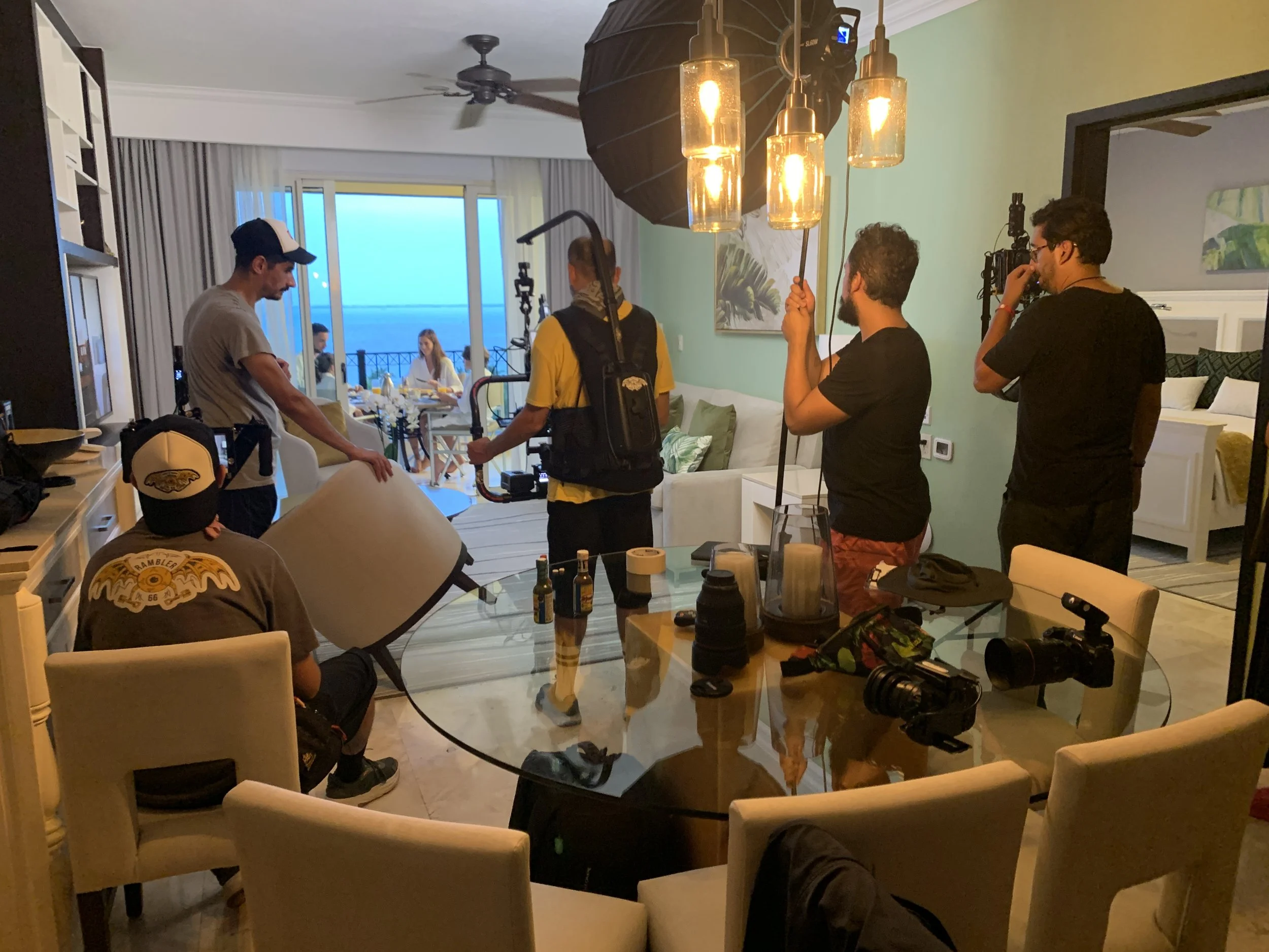 A film crew setting up equipment in a modern living room with a view of the ocean outside. Crew members are adjusting lights, cameras, and reflectors while preparing for a shoot near a dining table.