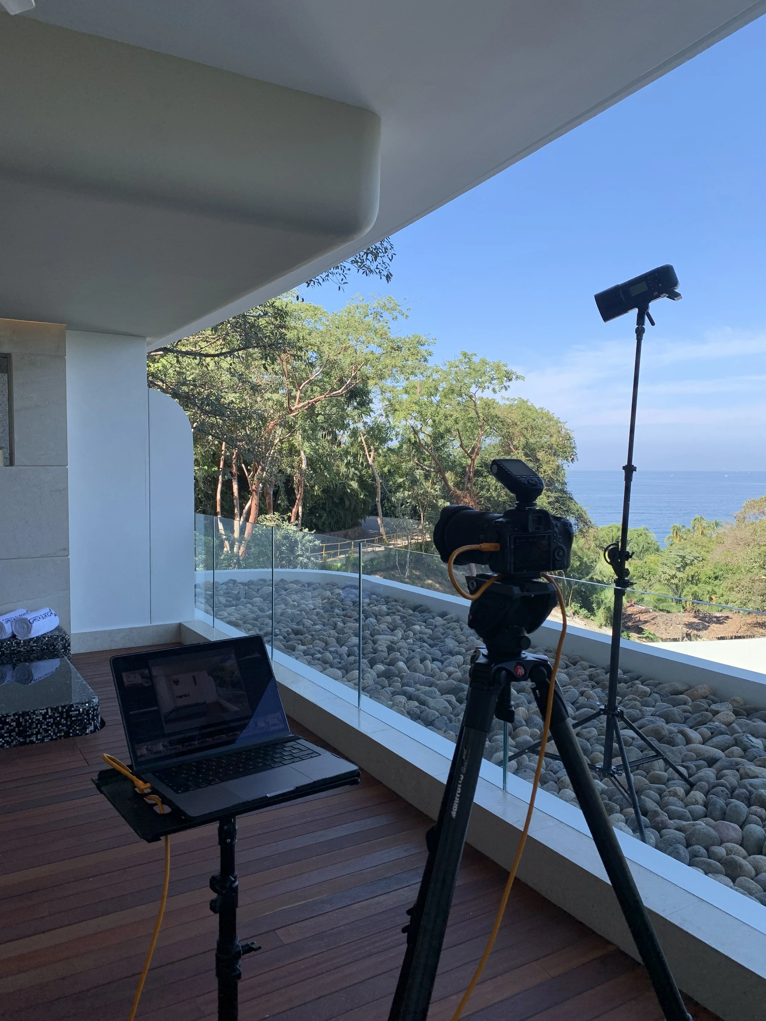 Photography setup on a balcony with a camera on tripod, laptop, and lighting equipment overlooking trees, rocks, and the ocean under a clear blue sky.