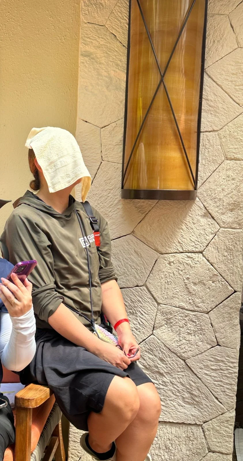Person sitting on a wooden bench with a towel on their head, wearing a hoodie and shorts, inside a room with stone walls and a tall, narrow decorative wall piece.