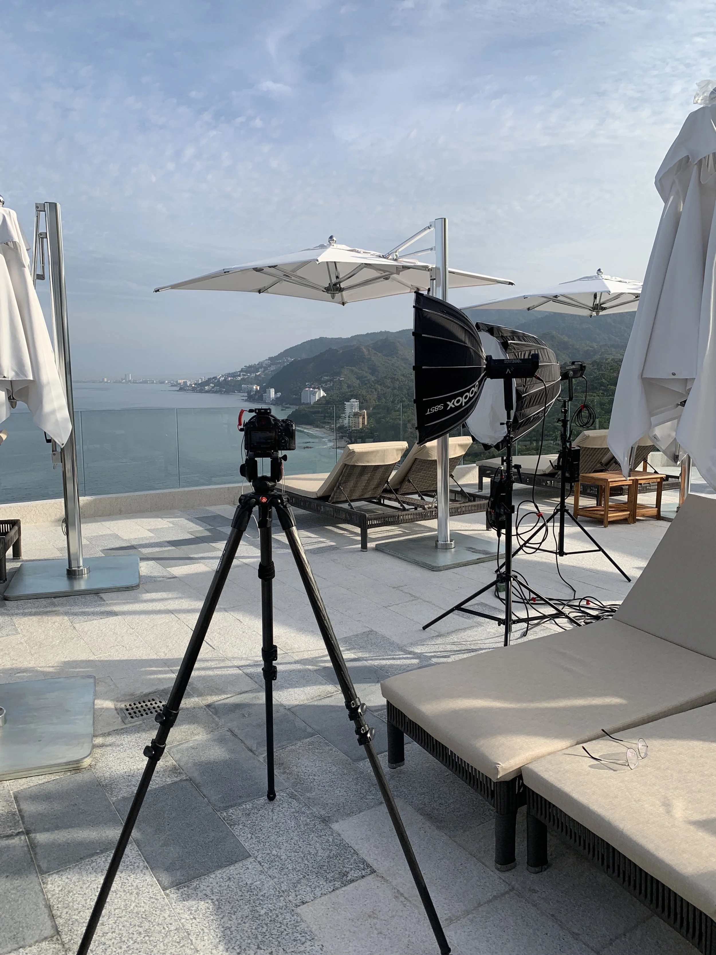 A rooftop photography or video shoot setup overlooking the water and hills. Equipment includes a camera on a tripod, studio lights, and white umbrellas for shade, with lounge chairs and a pair of glasses on a beige cushioned bench.