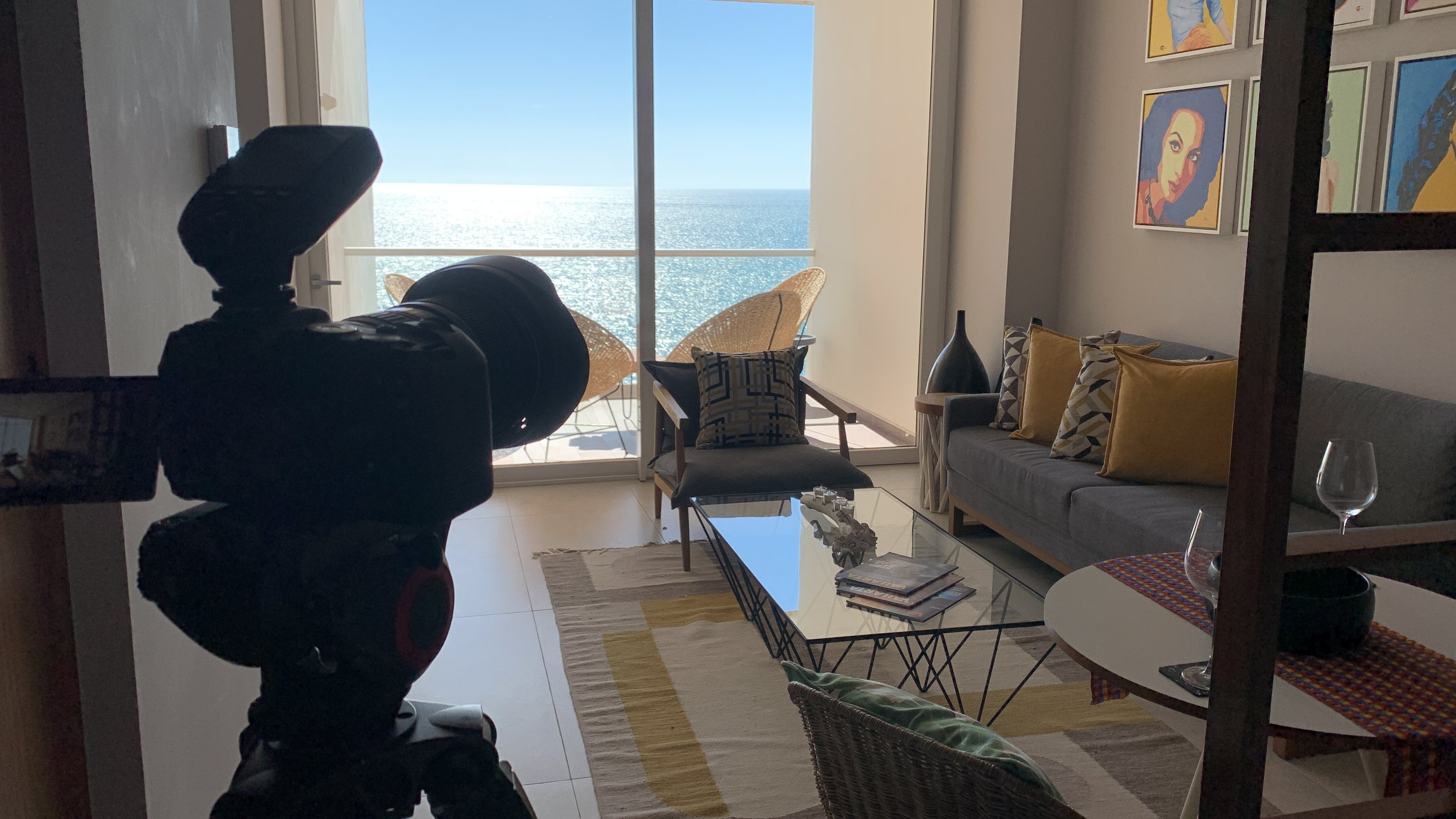 A living room with a sliding glass door opening to a balcony overlooking the ocean, a gray sofa with yellow and patterned pillows, a glass-top coffee table with magazines, a chair with a colorful pillow, and artwork on the walls.