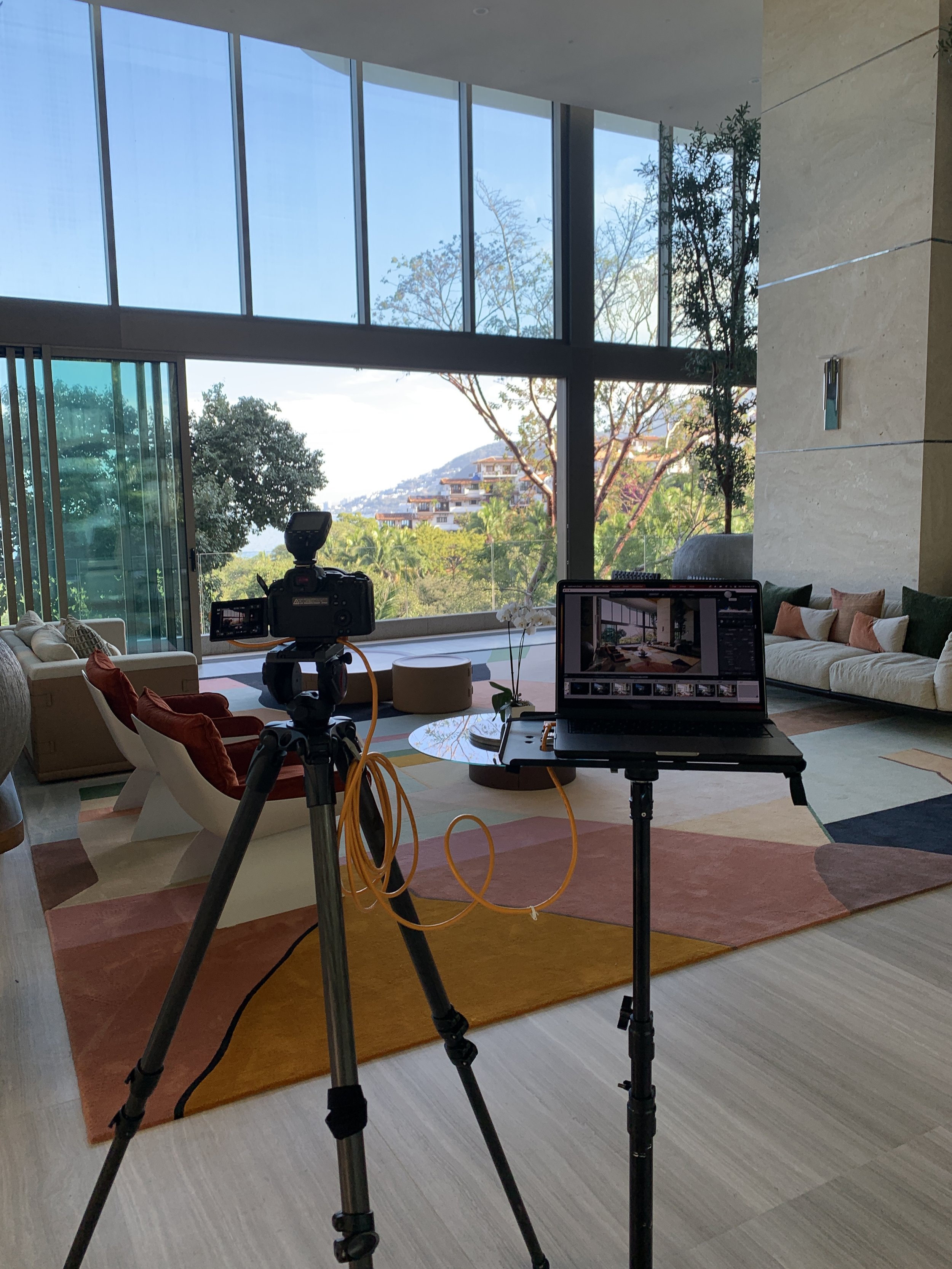 Interior shot of a modern living room with large glass windows overlooking a scenic outdoor view, featuring a camera and laptop set up on tripods for filming.