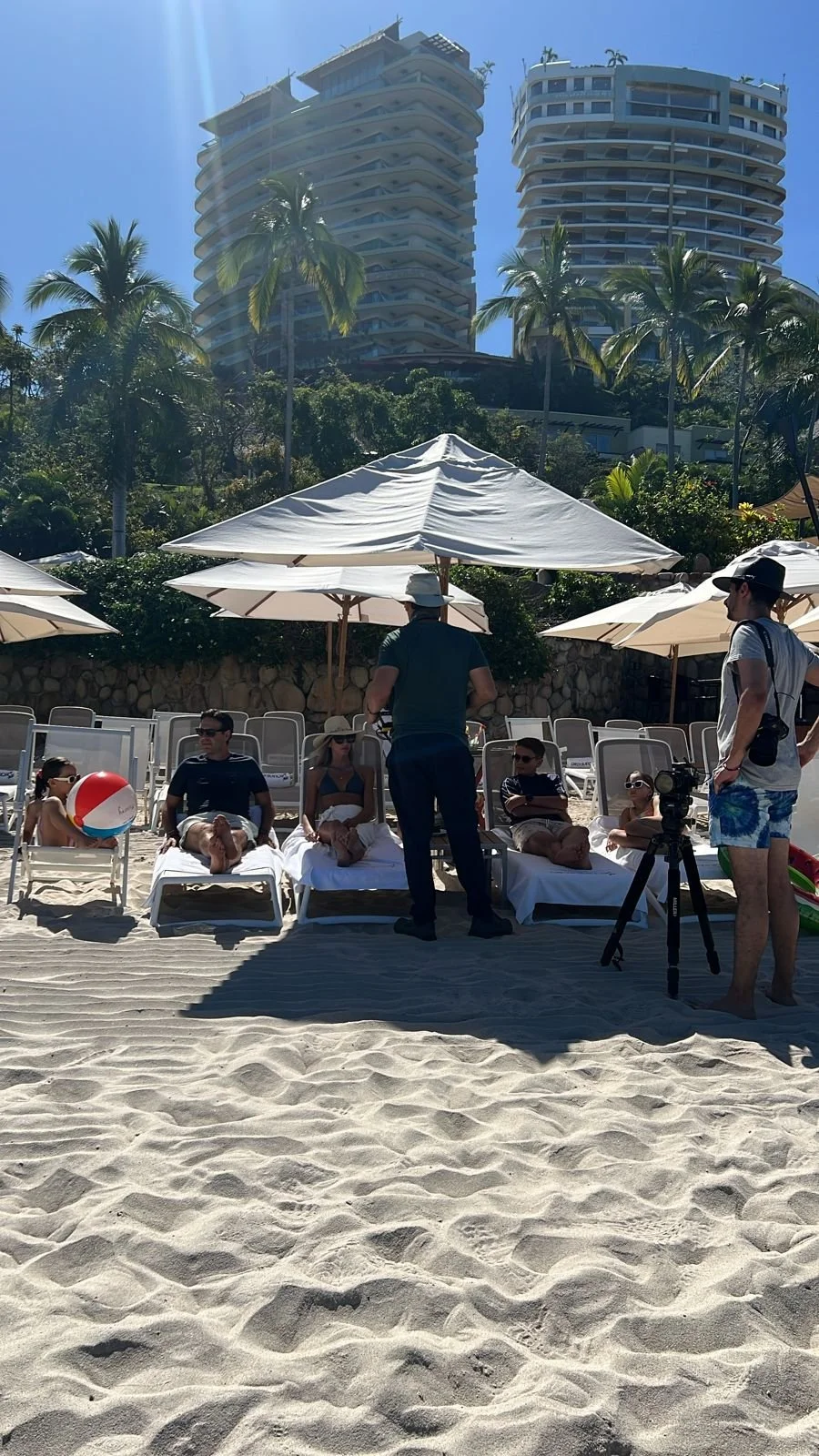 People relaxing on lounge chairs at a beach resort, with several palm trees, tall modern buildings, and beach umbrellas in the background on a sunny day.
