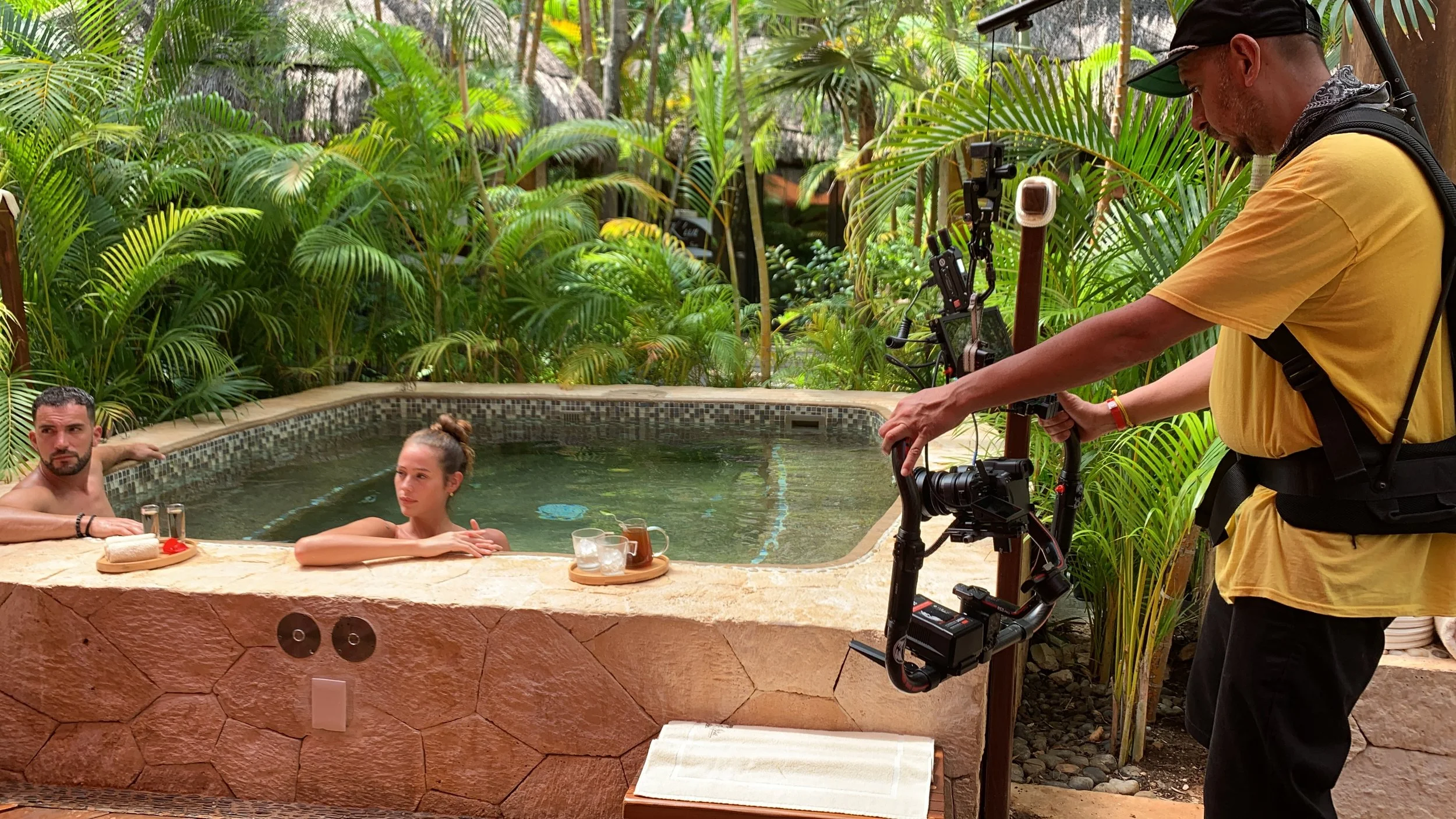A man and woman in a hot tub surrounded by lush green tropical plants, with a camera operator filming them using a stabilizer.