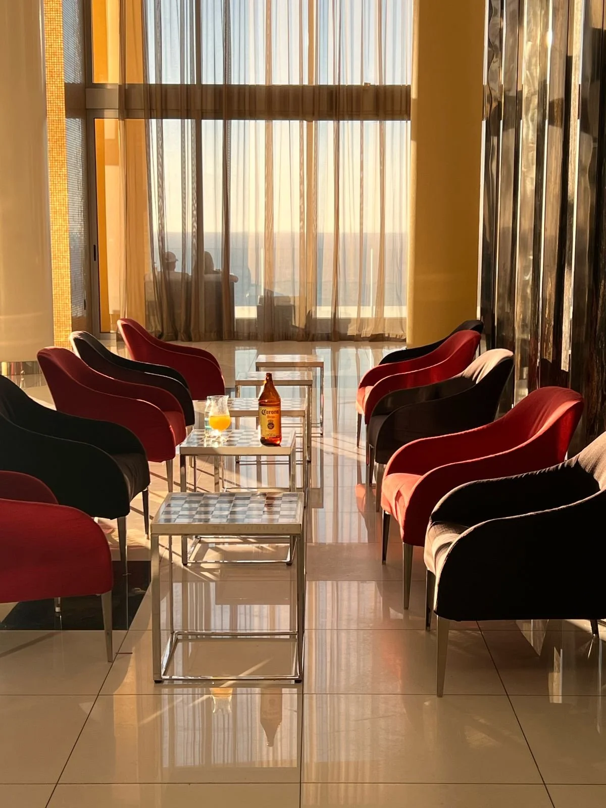 Sunlit hotel lounge with red and black chairs, glass tables, a bottle of Corona beer, a glass of orange juice, and sheer curtains overlooking the ocean at sunset.