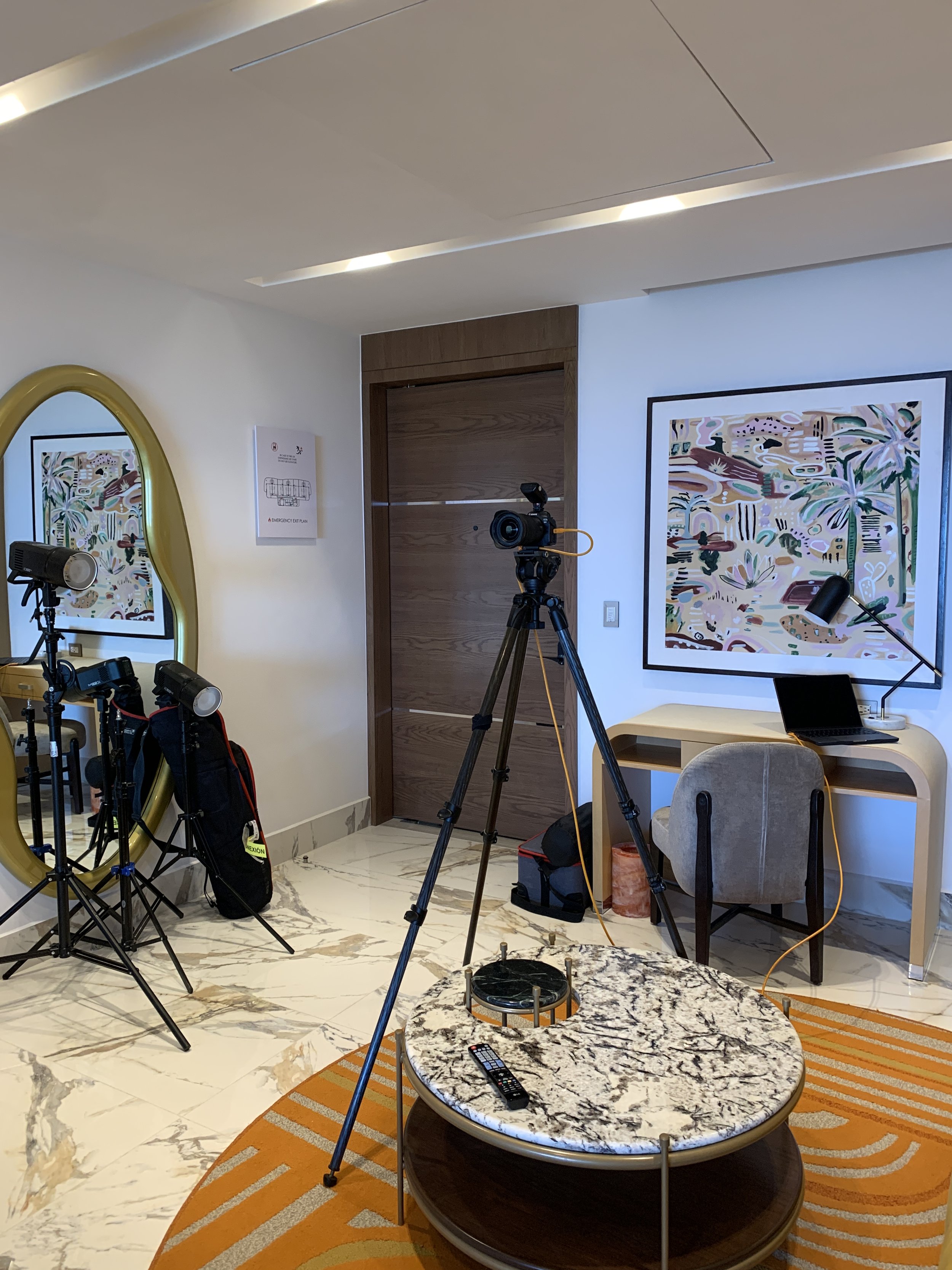 A hotel room or conference space with cameras and equipment set up for filming or photography. Includes a round marble table with a TV remote, a laptop on a wooden desk, a beige chair, framed artwork on the wall, a large mirror with a yellow frame, and a wooden door.