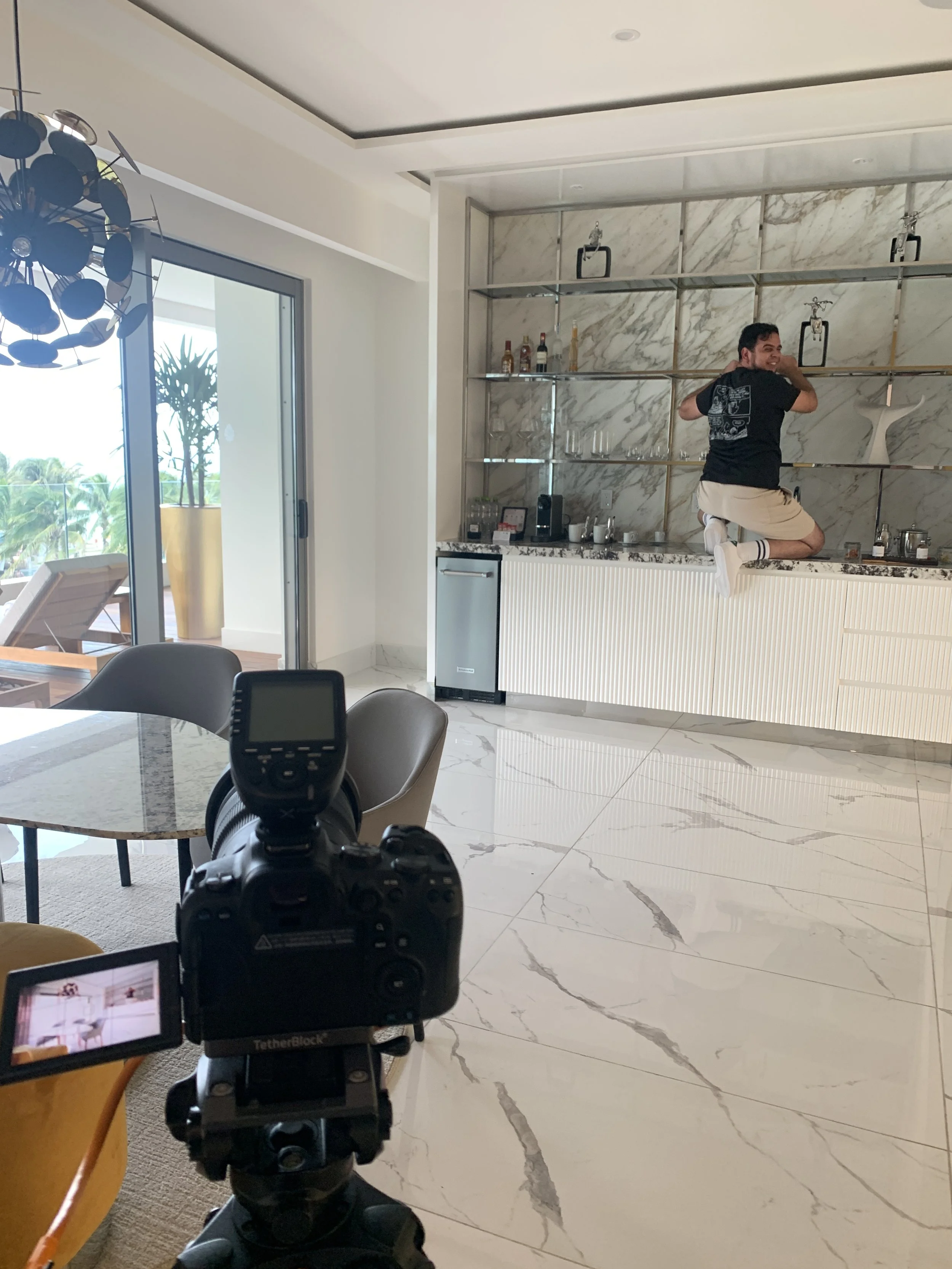 A person kneeling on a kitchen counter with a camera set up in the foreground. The kitchen has marble walls and floor, with a sliding glass door leading to an outdoor patio with a lounge chair and greenery.