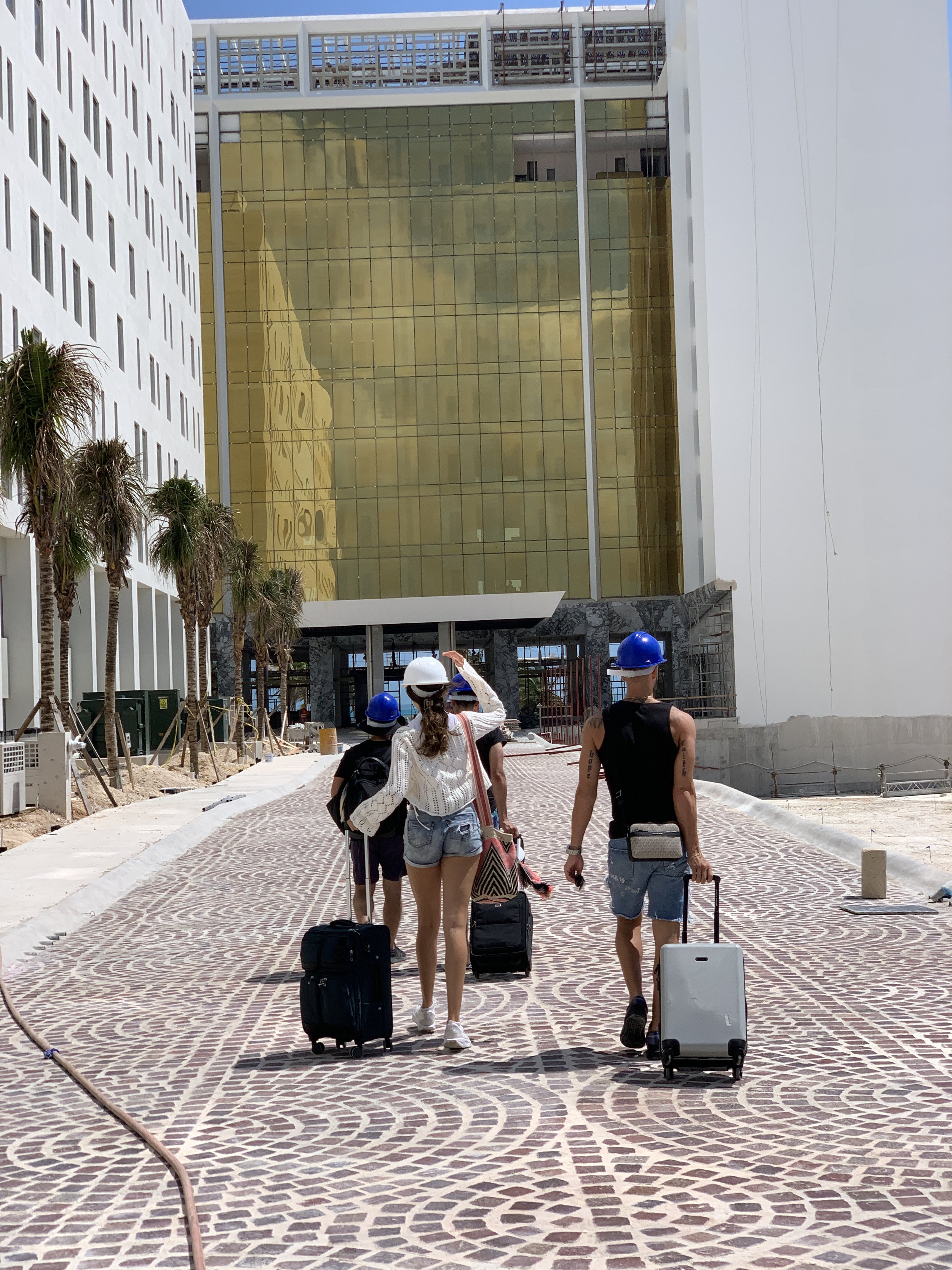 Group of tourists walking with luggage outside modern white building with golden glass facade, palm trees lining sidewalk