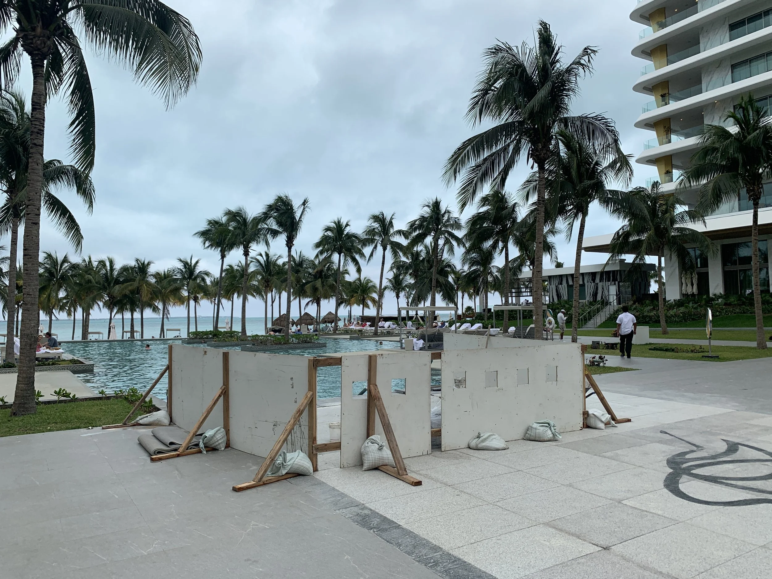 Construction barrier near a swimming pool at a hotel with palm trees, lounge chairs, and a view of the ocean in the background.