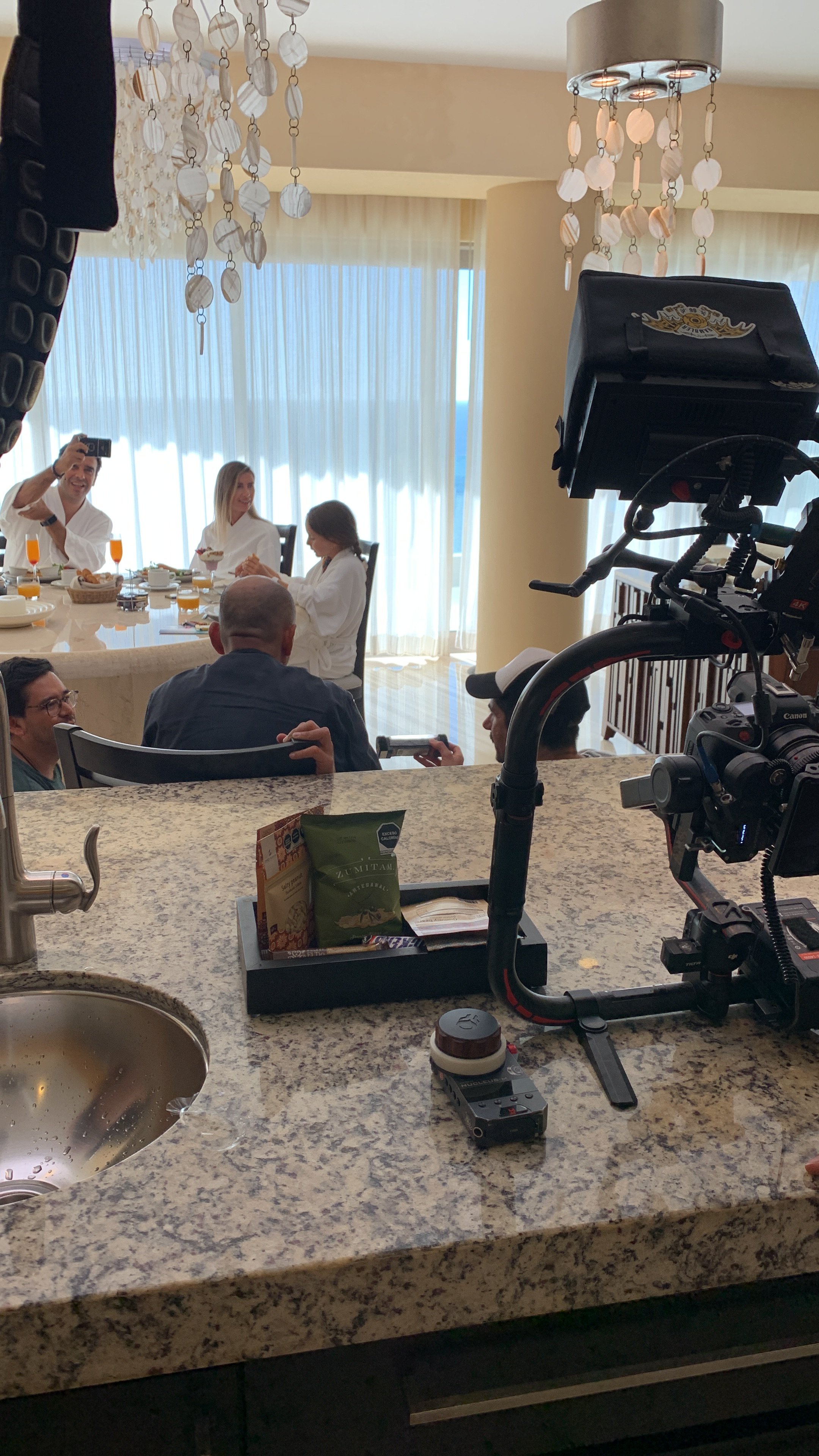 People gathered around a dining table with breakfast items, some taking photos, in a well-lit room with large windows and decorative hanging chandeliers.