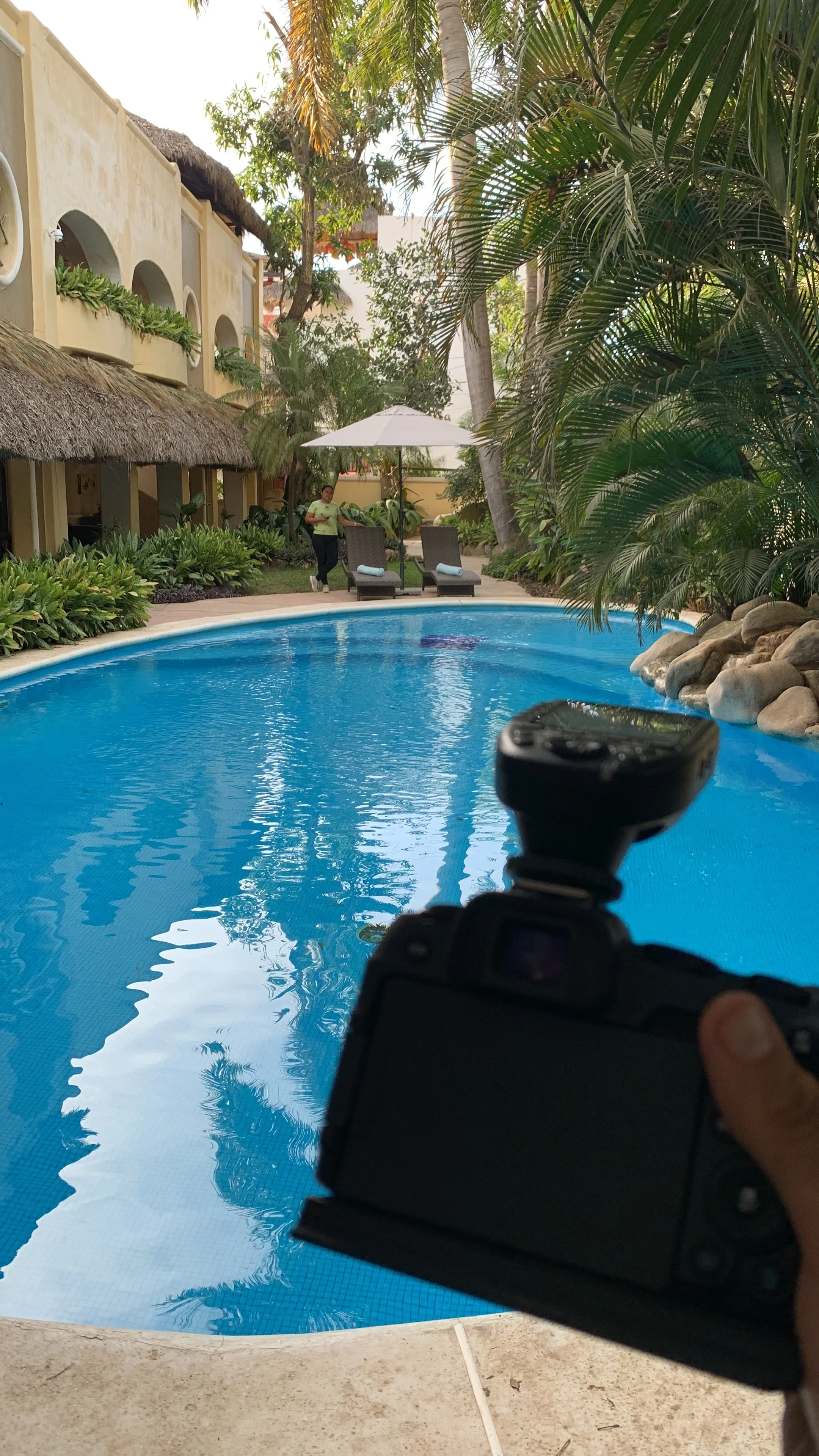 A swimming pool surrounded by tropical plants and lounge chairs, with a person walking near the chairs and photographer's hand holding a camera in the foreground.