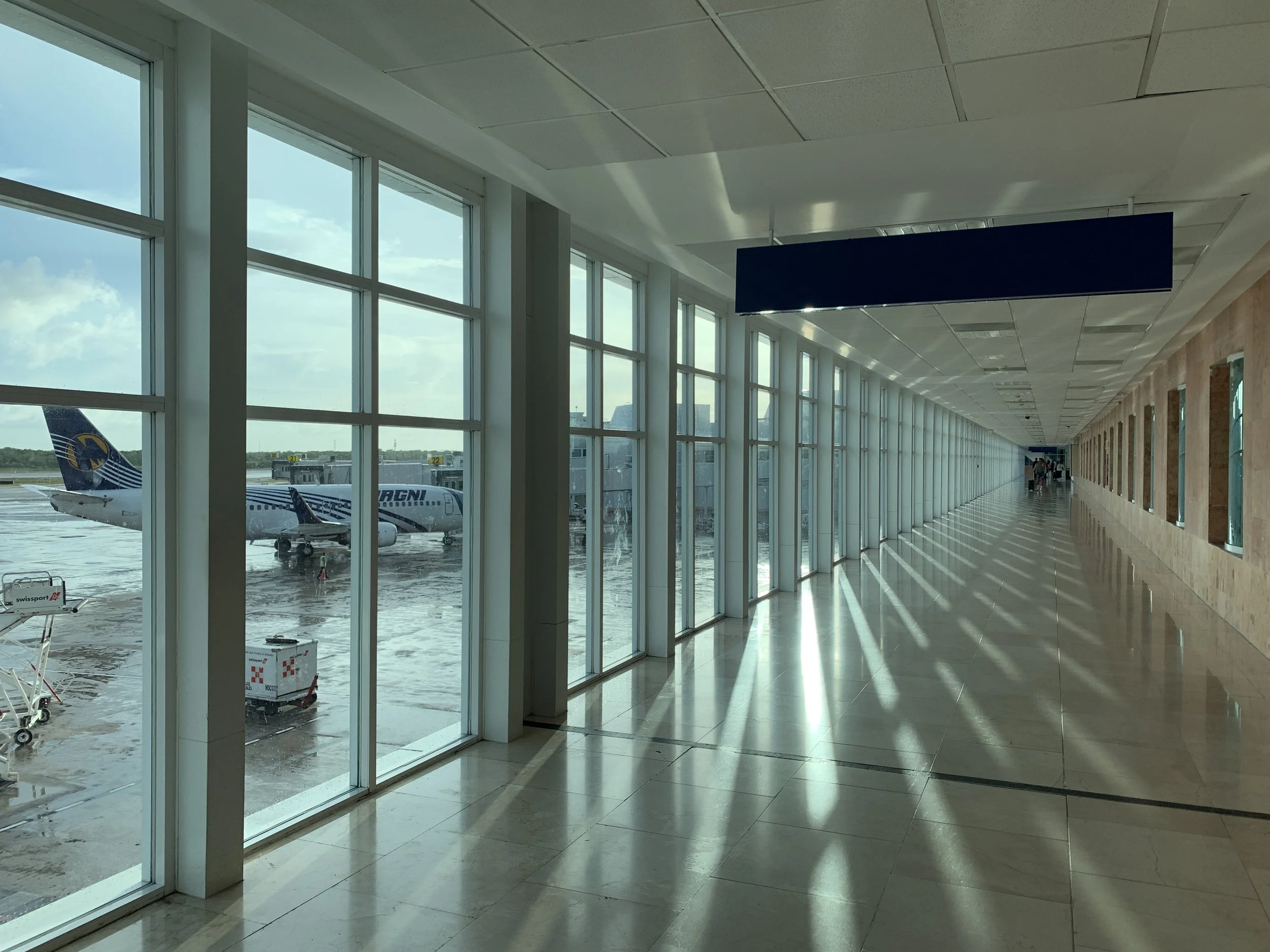 Empty airport terminal corridor with large windows showing airplanes outside, sunlight casting shadows on the floor.