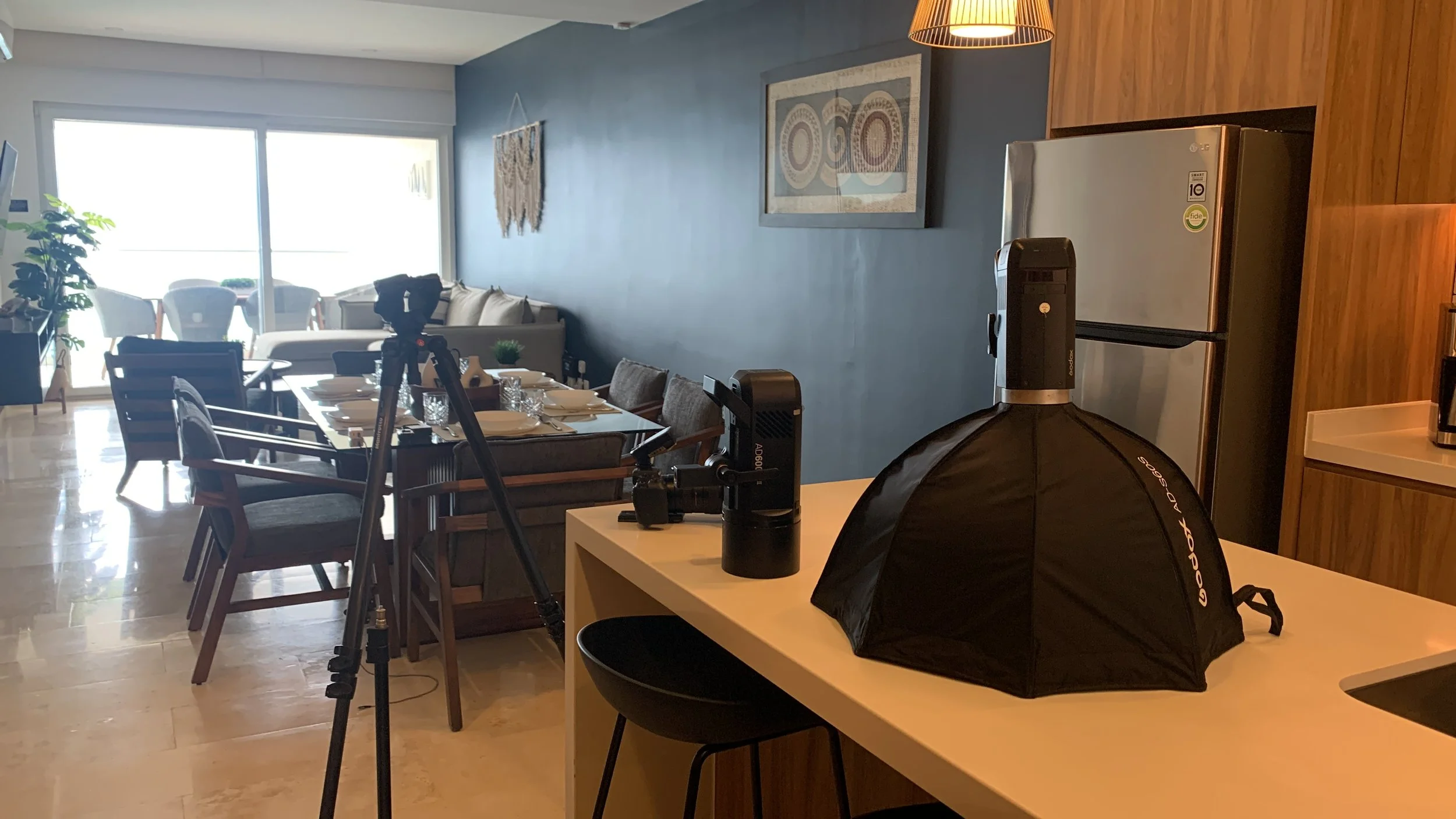 A living room and kitchen area set up for filming or photography, with a camera on a tripod, lighting equipment, a dining table with plates and glasses, a sofa, chairs, a window with an outdoor view, a refrigerator, and wall art.