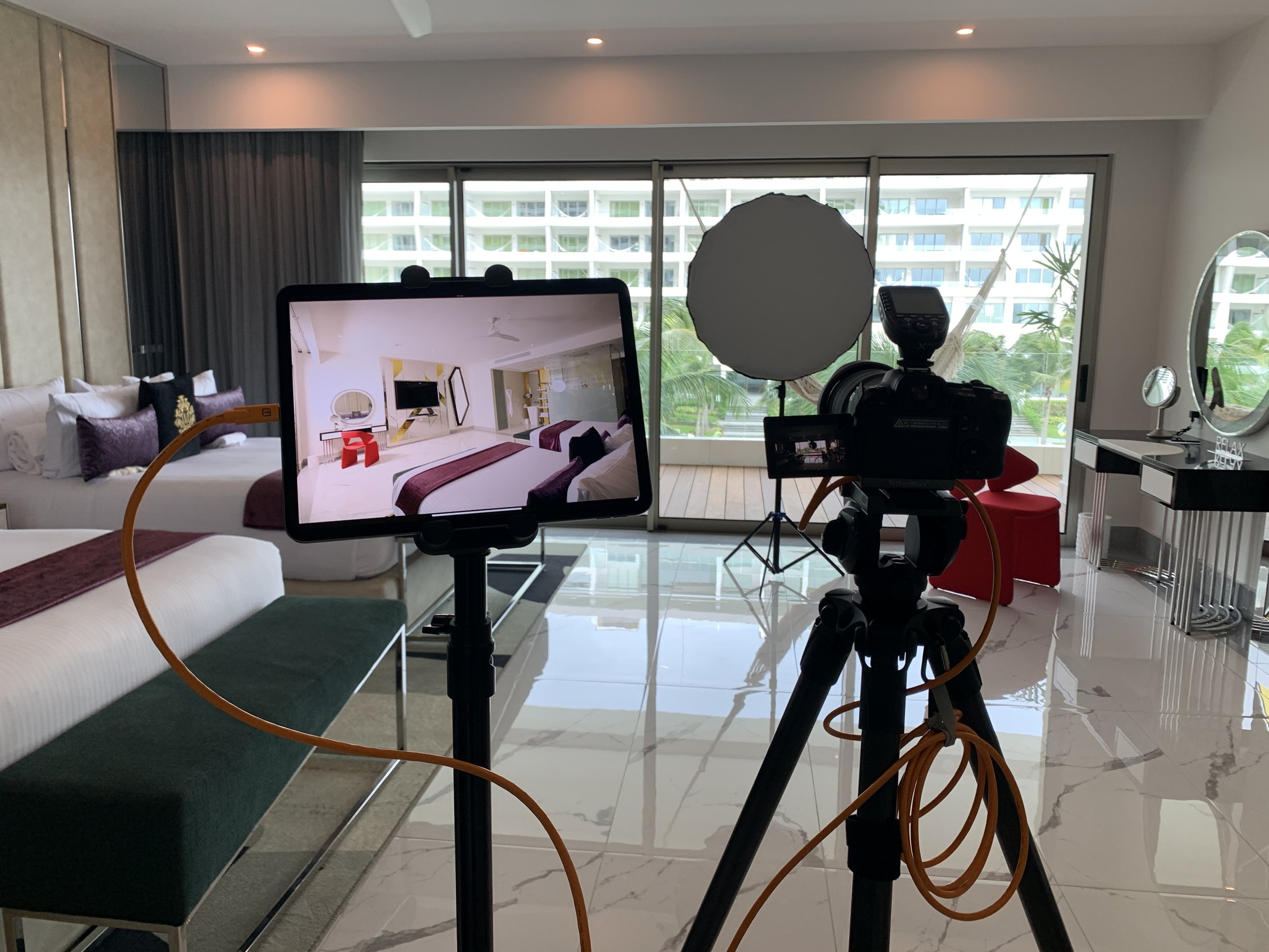 A filming setup inside a modern hotel room with two beds, a large window with a view of other buildings and palm trees, a tripod with a camera, a monitor, a soft light, a red chair, and a vanity table with mirror.