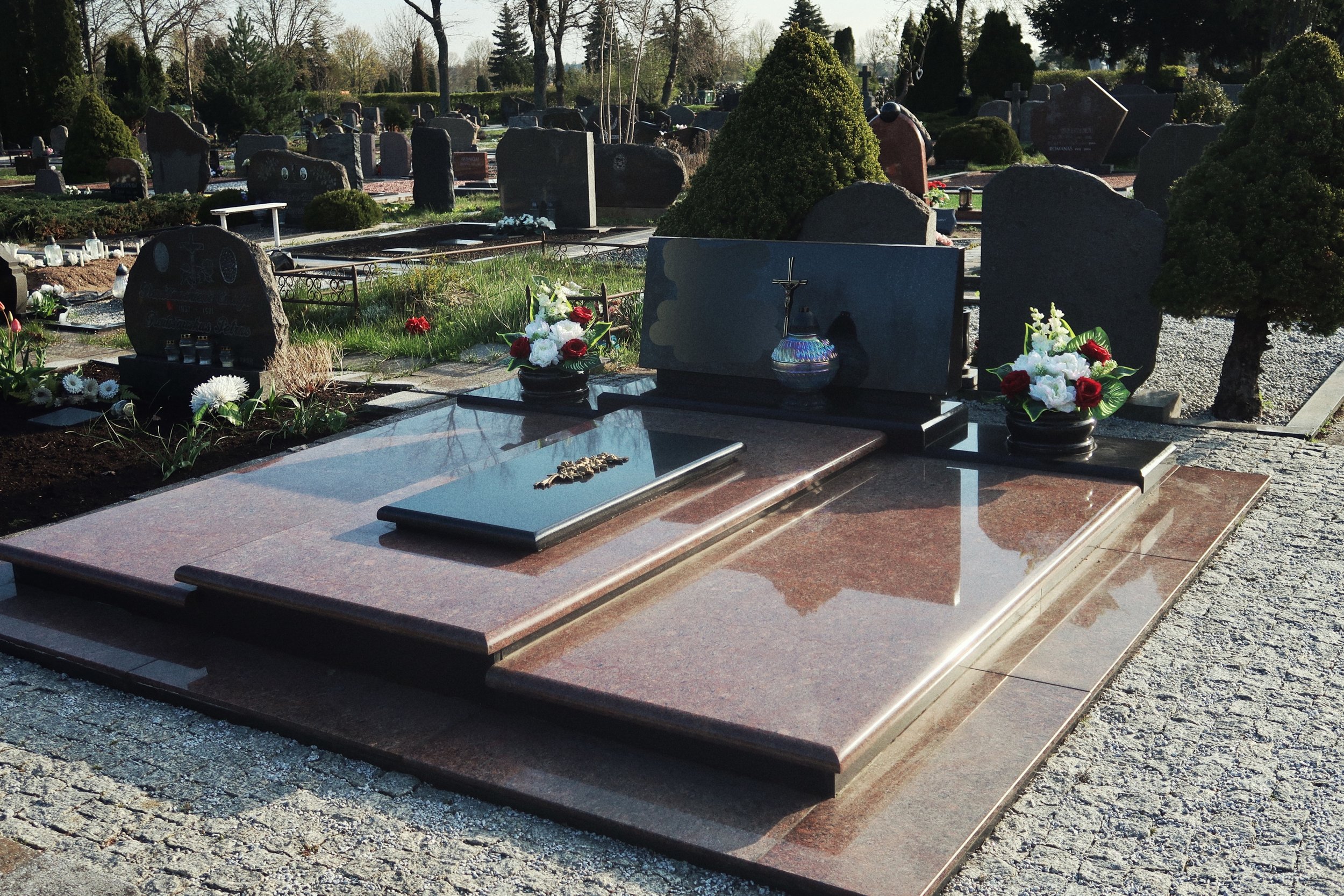 Gravestone on polished brown granite slab with crucifix, flowers, and urn