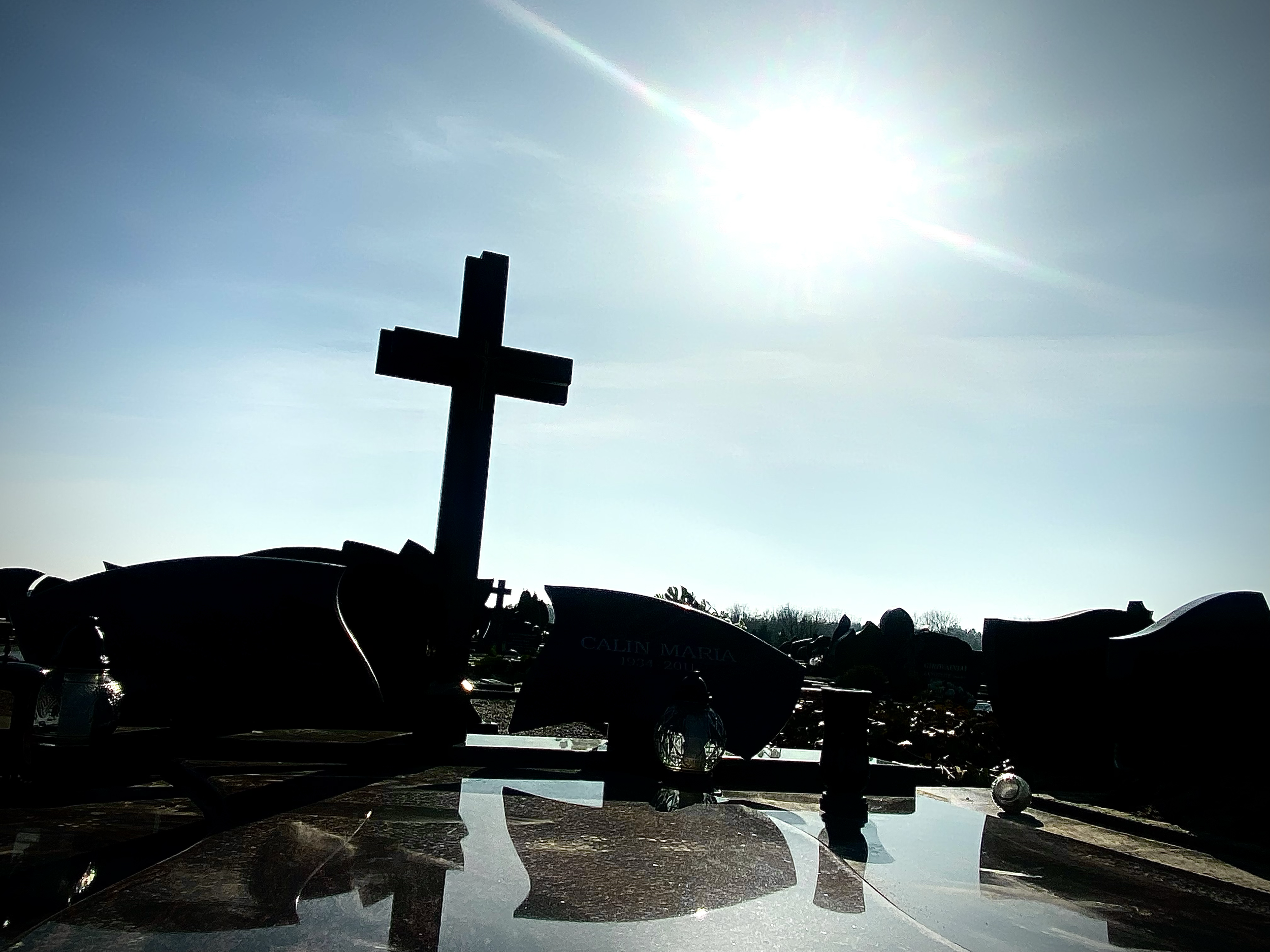 Silhouette of a cross at a cemetery with gravestones, under a bright sunlit sky.