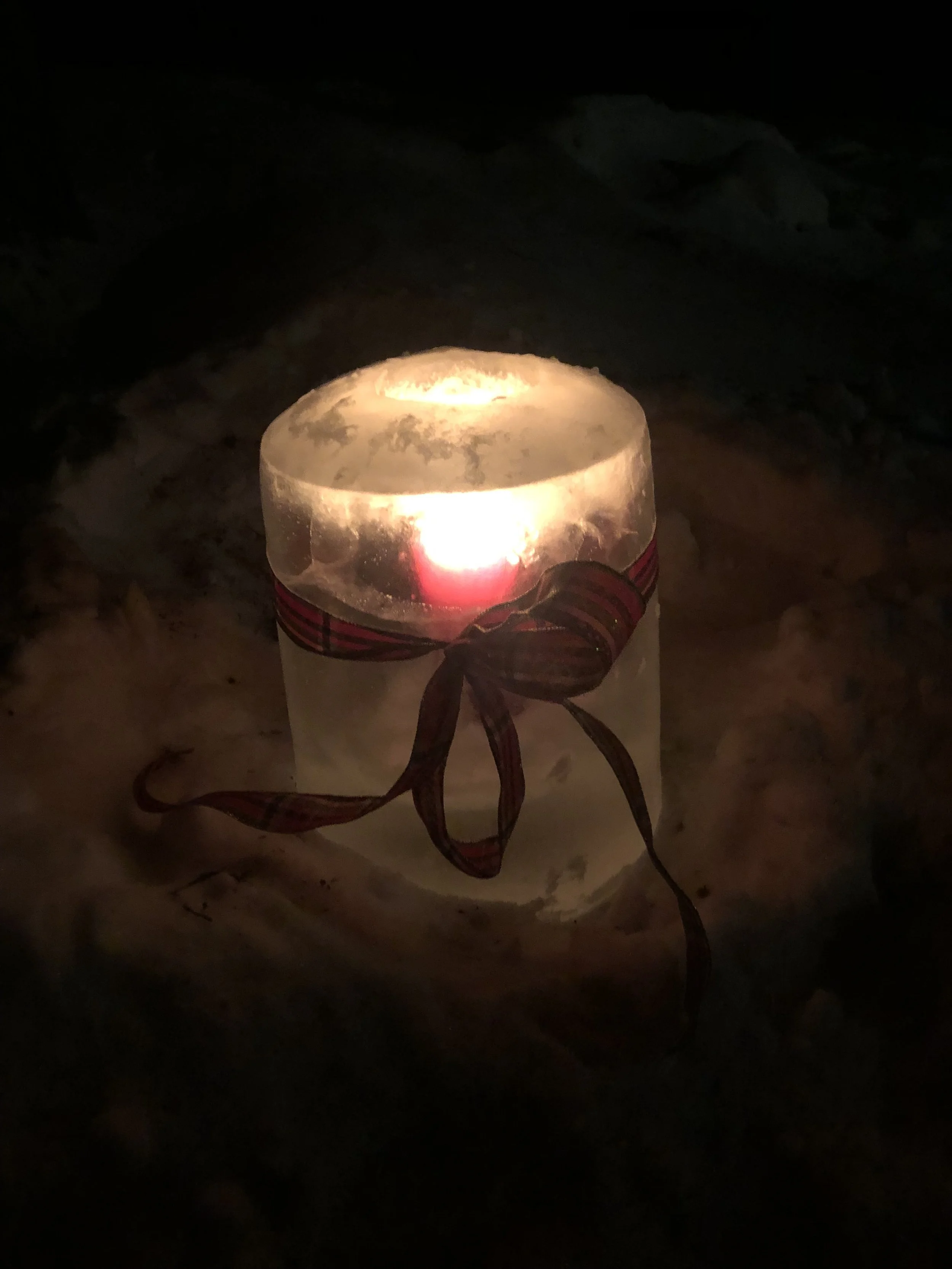 A lit candle in an ice holder with a plaid ribbon tied around it, placed on snow at night.