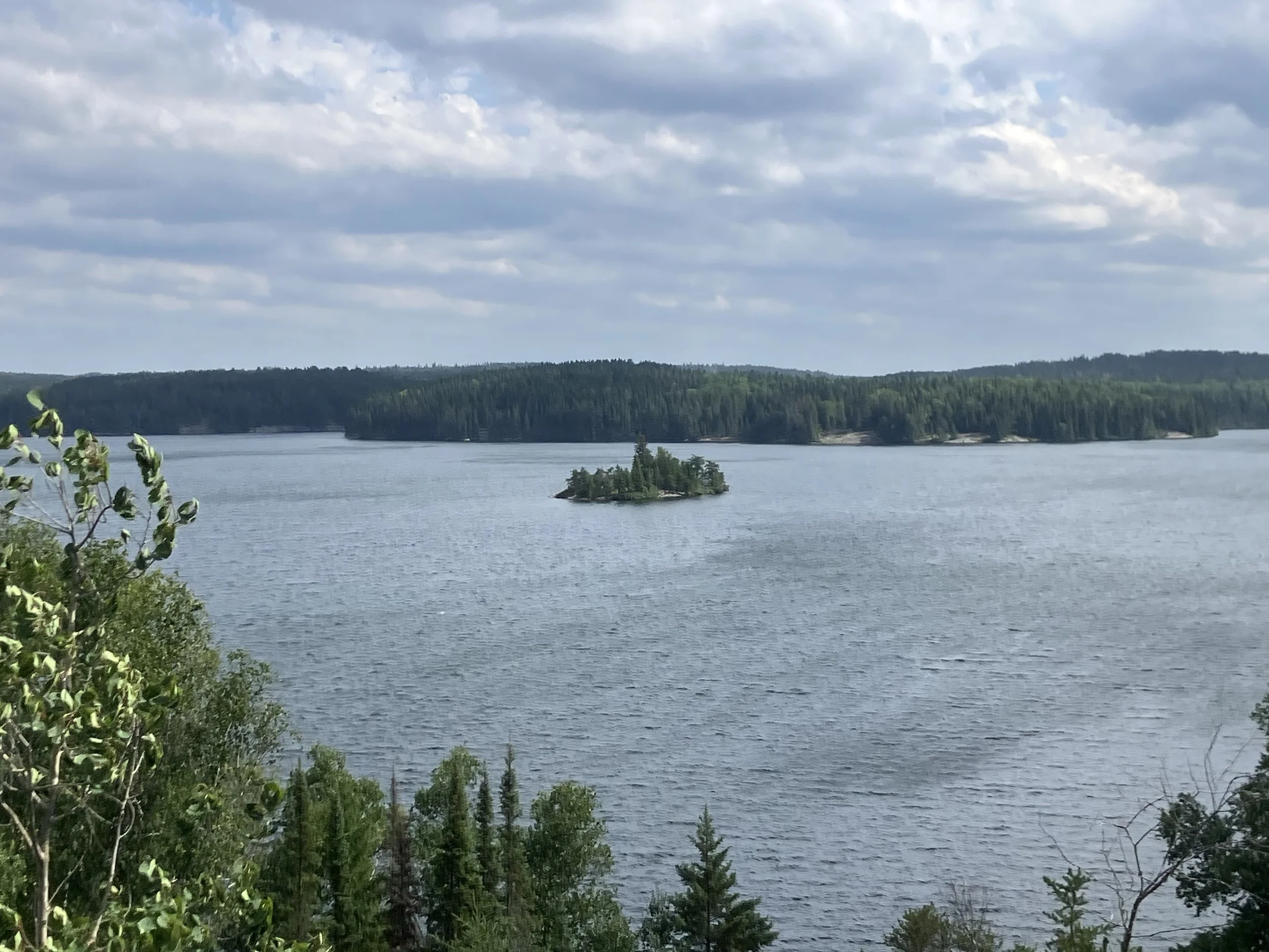 a view from above of a lake with blue sky, grey clouds and green trees on the horizon