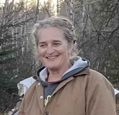 A person wearing a brown jacket and a gray hoodie, smiling outdoors with trees in the background.