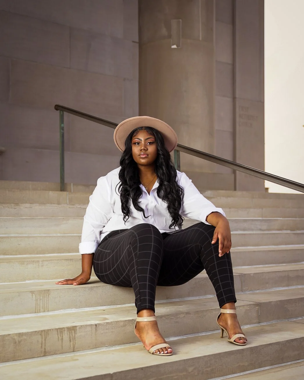 A woman with long black curly hair, wearing a beige hat, white shirt, black checkered pants, and beige high heels, sitting on steps outside a building with stone walls and a railing.