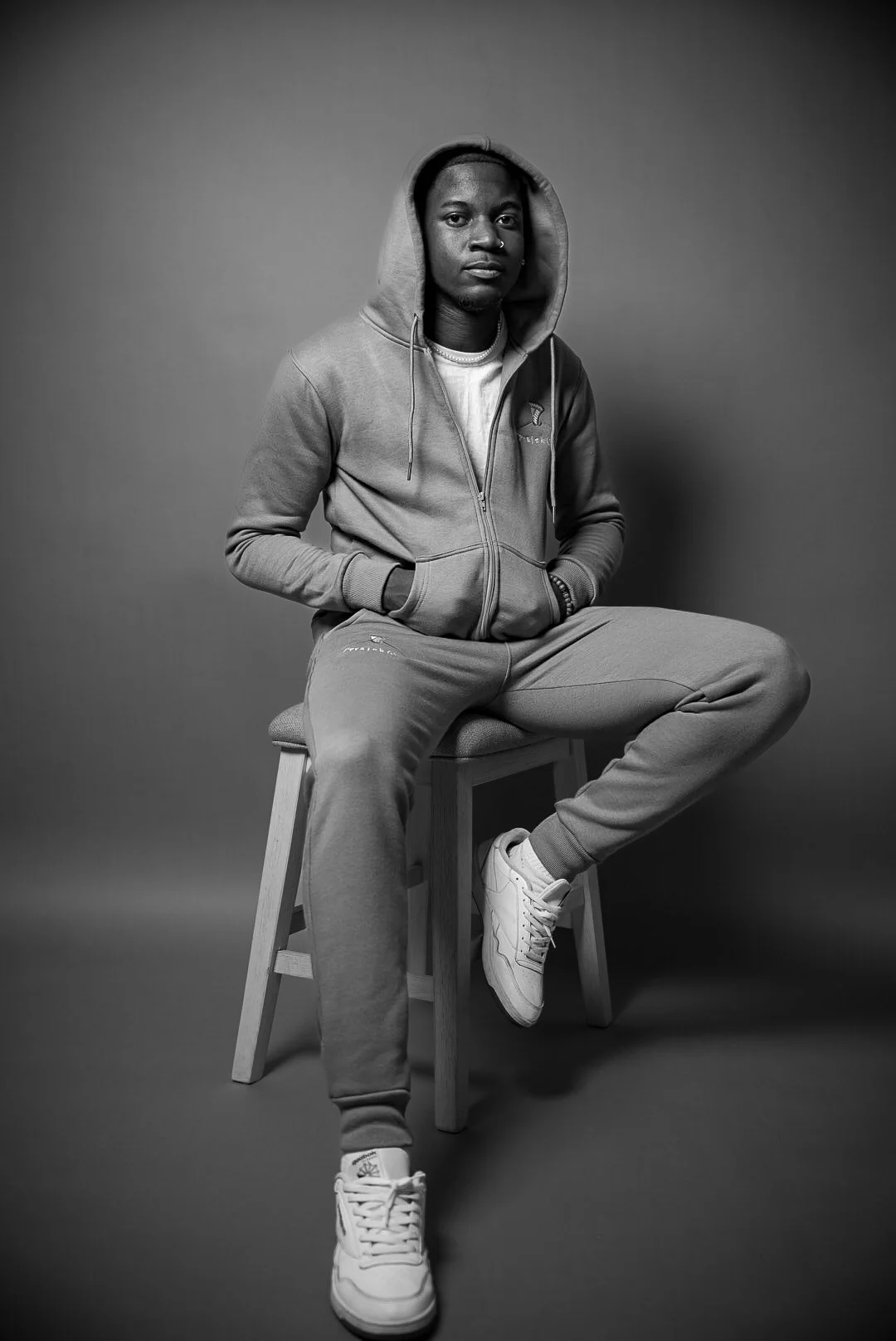 Black and white photo of a young man sitting on a stool, wearing a hoodie, sweatpants, and sneakers, in a studio setting