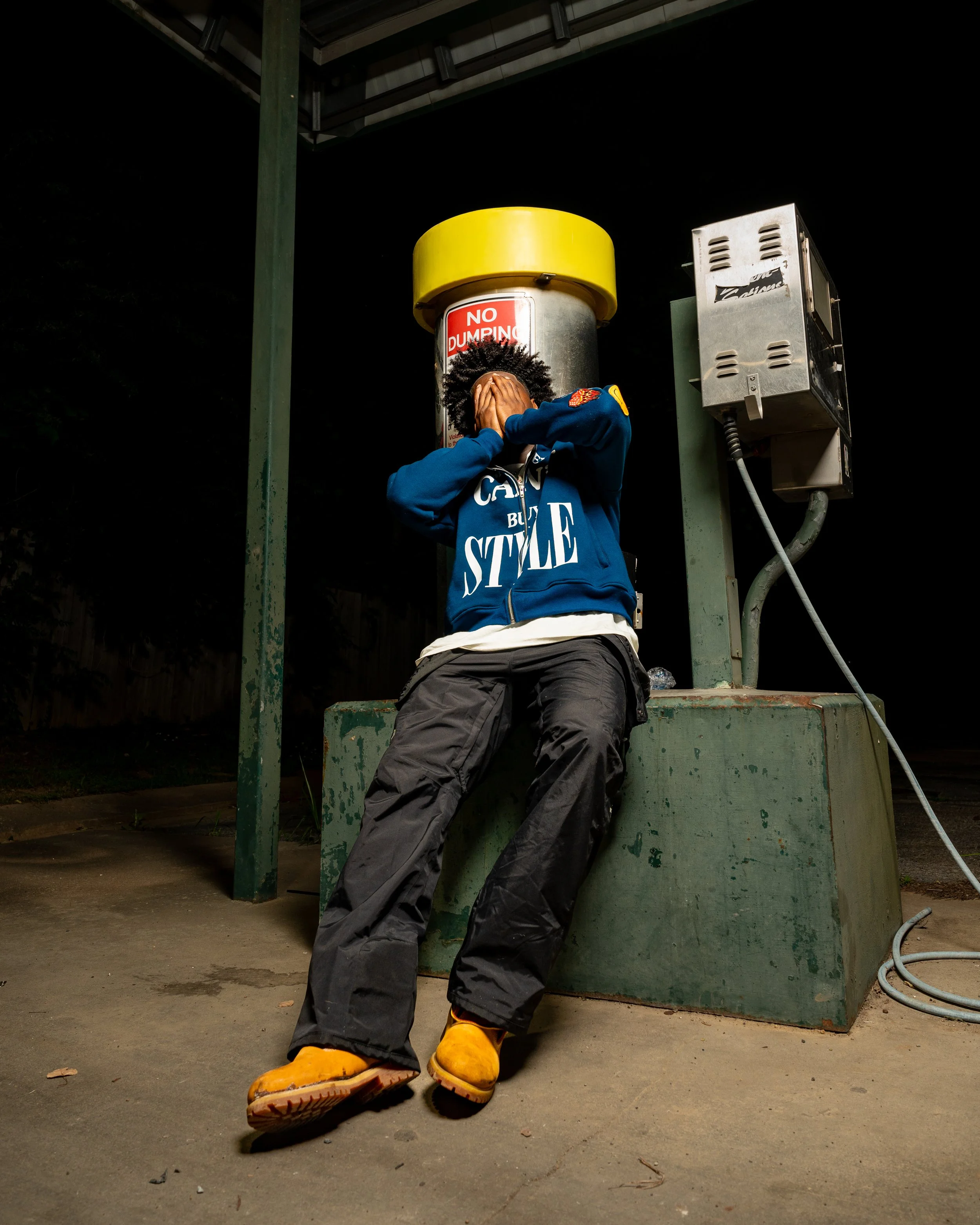 Person sitting on a green concrete platform with hands covering face, wearing a blue hoodie with text, black pants, and yellow shoes at night.