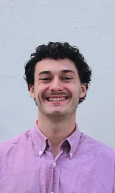 Ein lachender junger Mann mit dunklen, lockigen Haaren trägt ein lila Hemd vor einer hellgrauen Wand.