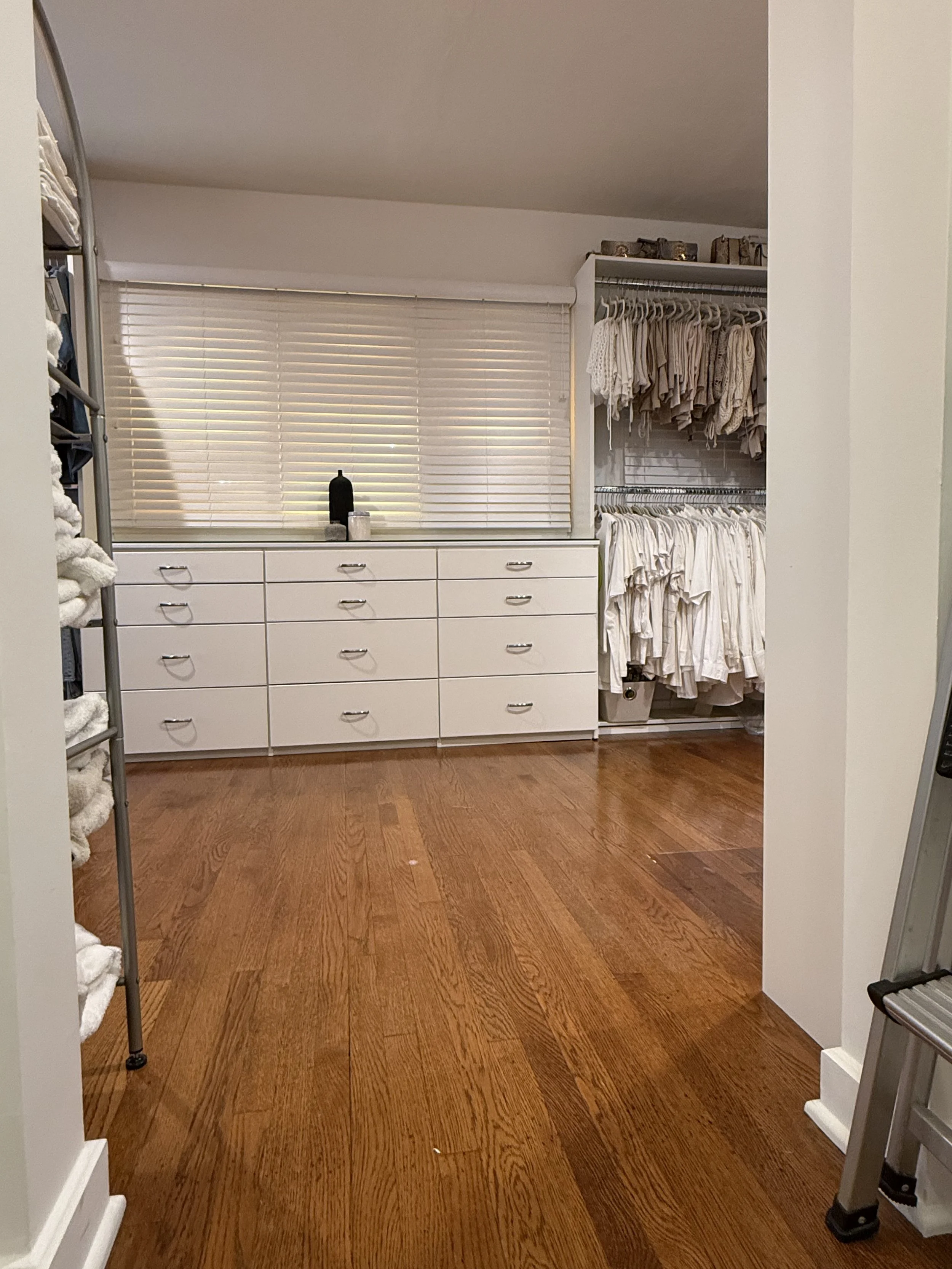 View of a walk-in closet with white drawers, hanging clothing in neutral colors, and a window with closed blinds. Wooden floor and some storage boxes on a shelf.