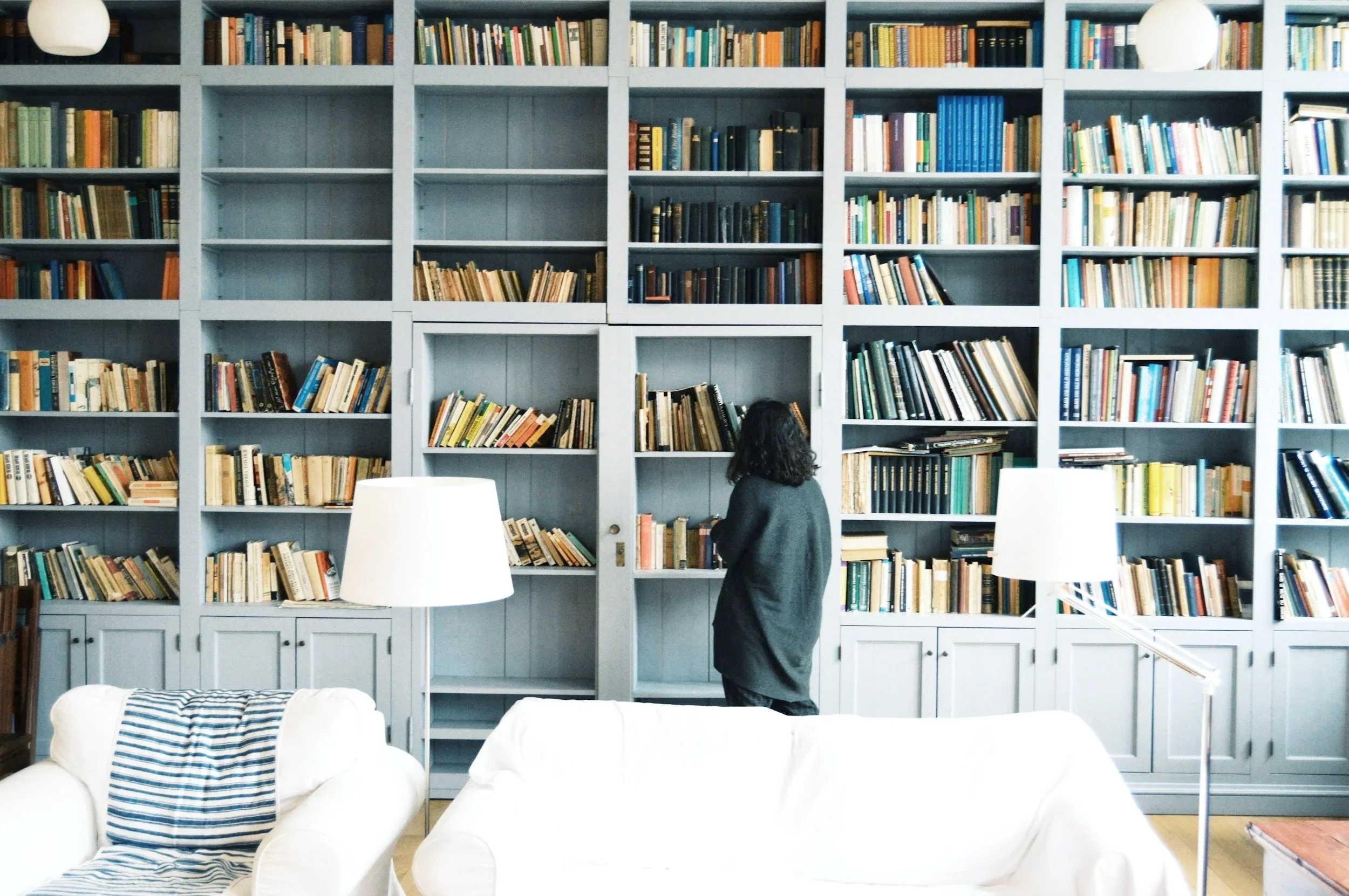 A woman with curly hair wearing a gray sweater stands in front of a large bookshelf filled with numerous books in a living room or library. White sofas and lamps are visible in the foreground.