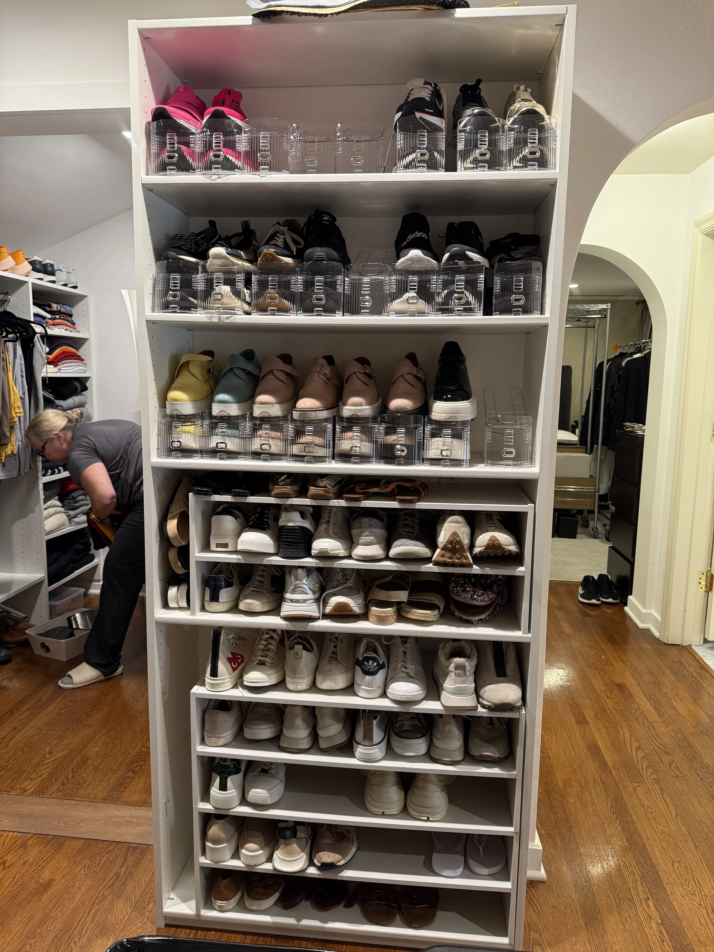 White bookshelf with multiple pairs of sneakers on each shelf, in a room with wooden flooring and gray walls.