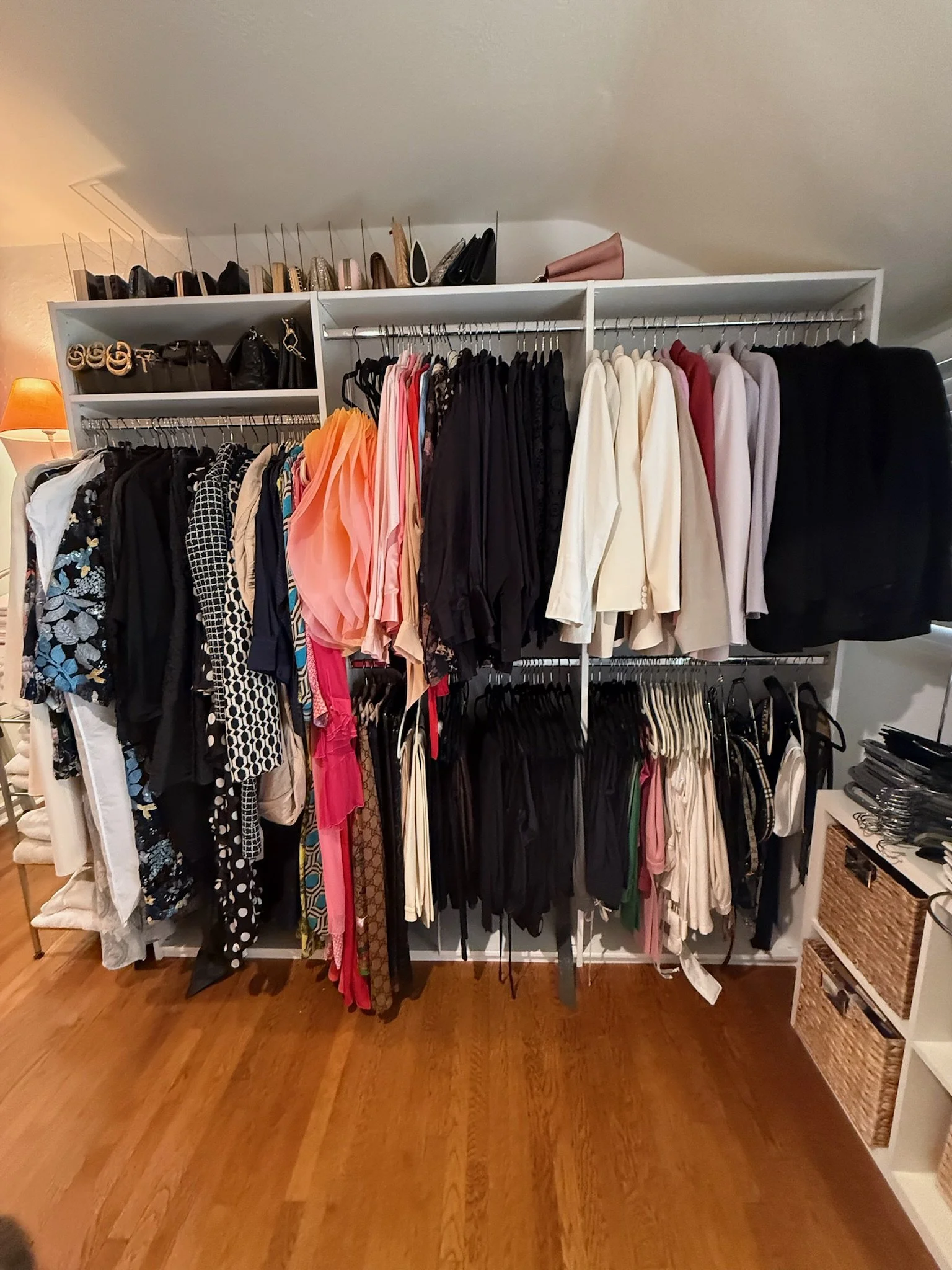 A large white closet filled with hanging clothes, bags, and shoes on top, situated on a hardwood floor in a room with beige walls and a small chest of drawers on the right.
