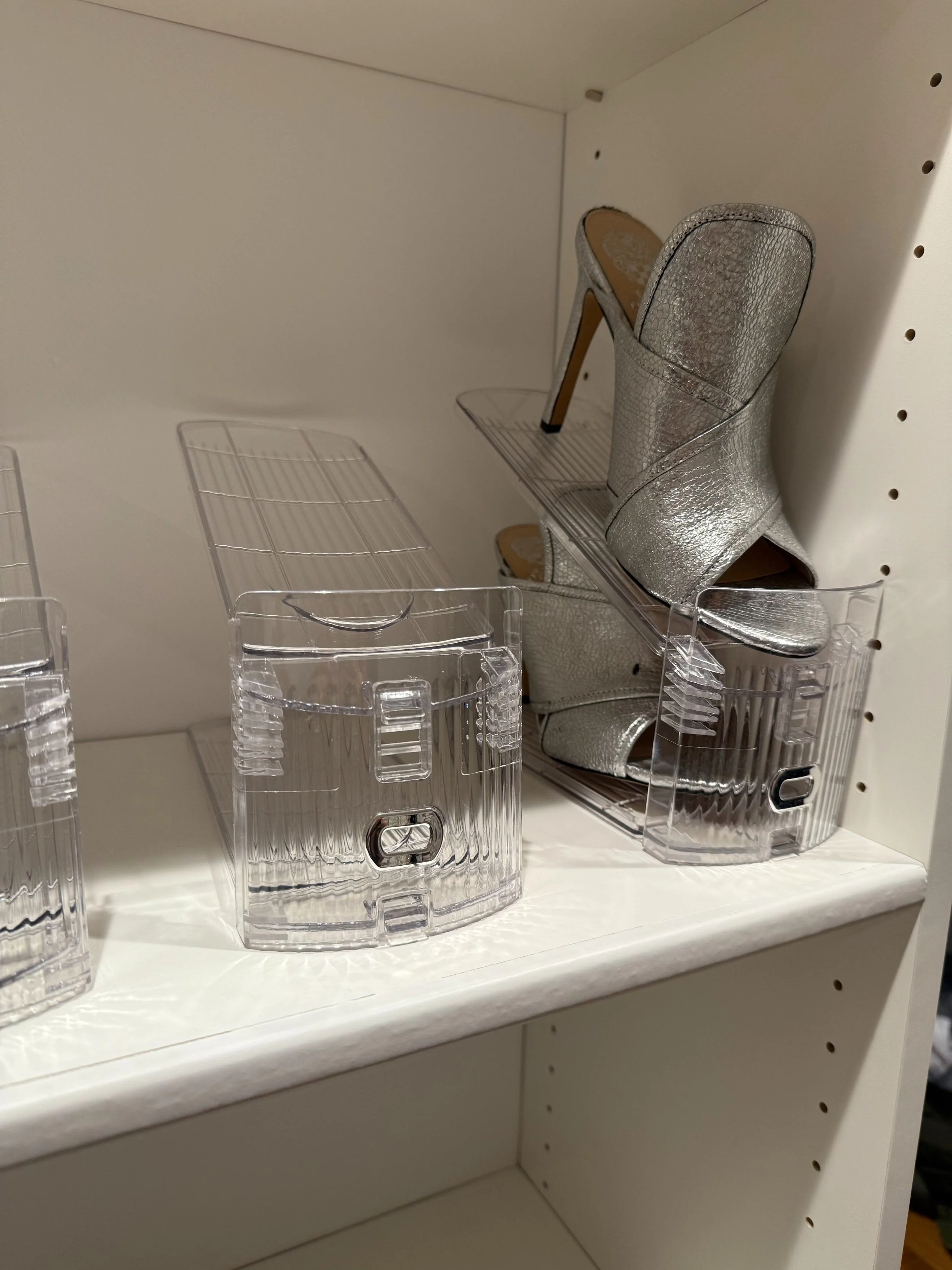 A pair of shiny silver high-heeled shoes on a white shelf, with some empty clear plastic containers nearby.