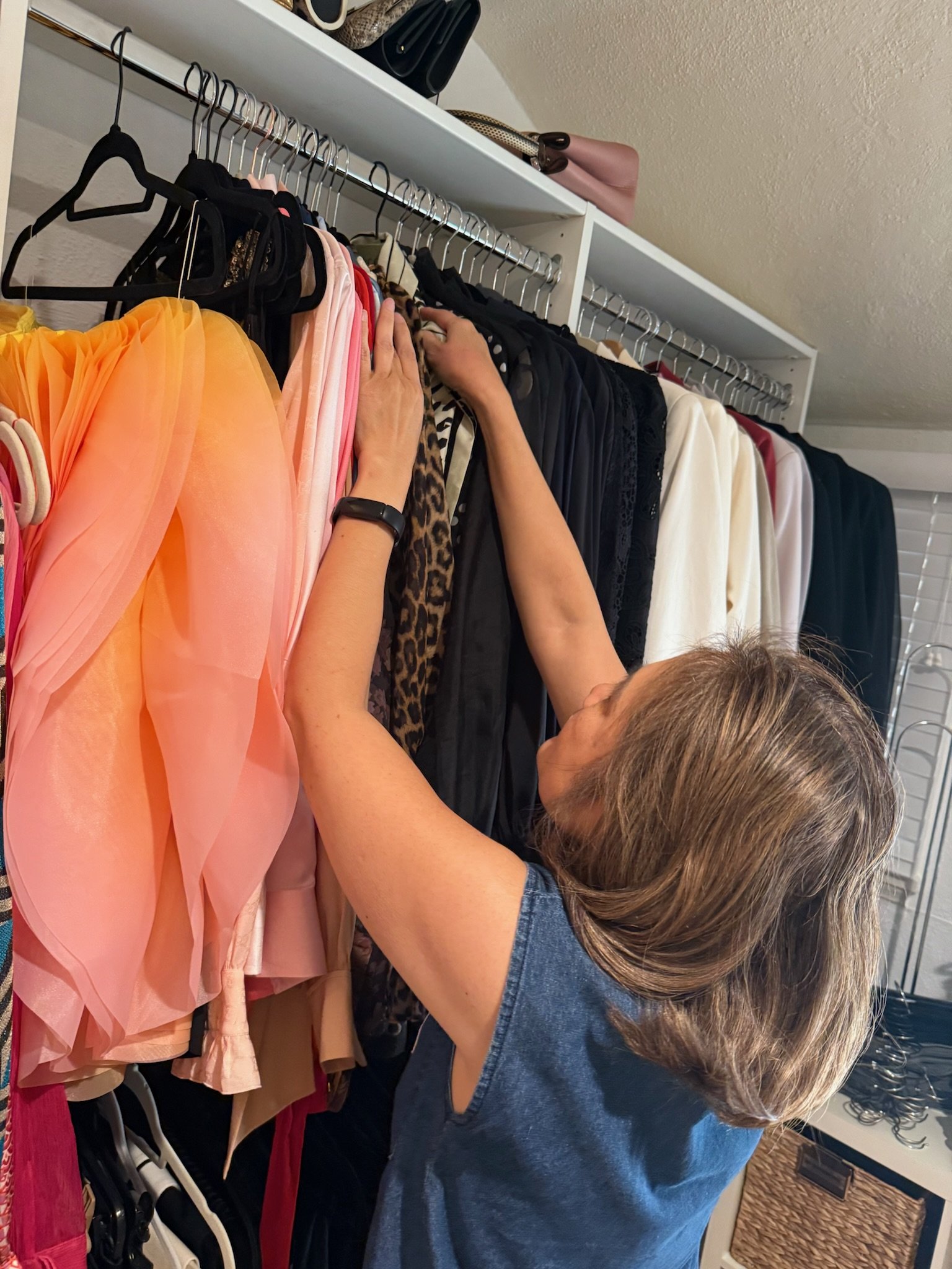 A woman shopping for clothes on a clothing rack, looking at various dresses and tops in different colors and patterns. She is wearing a blue shirt and a black watch.