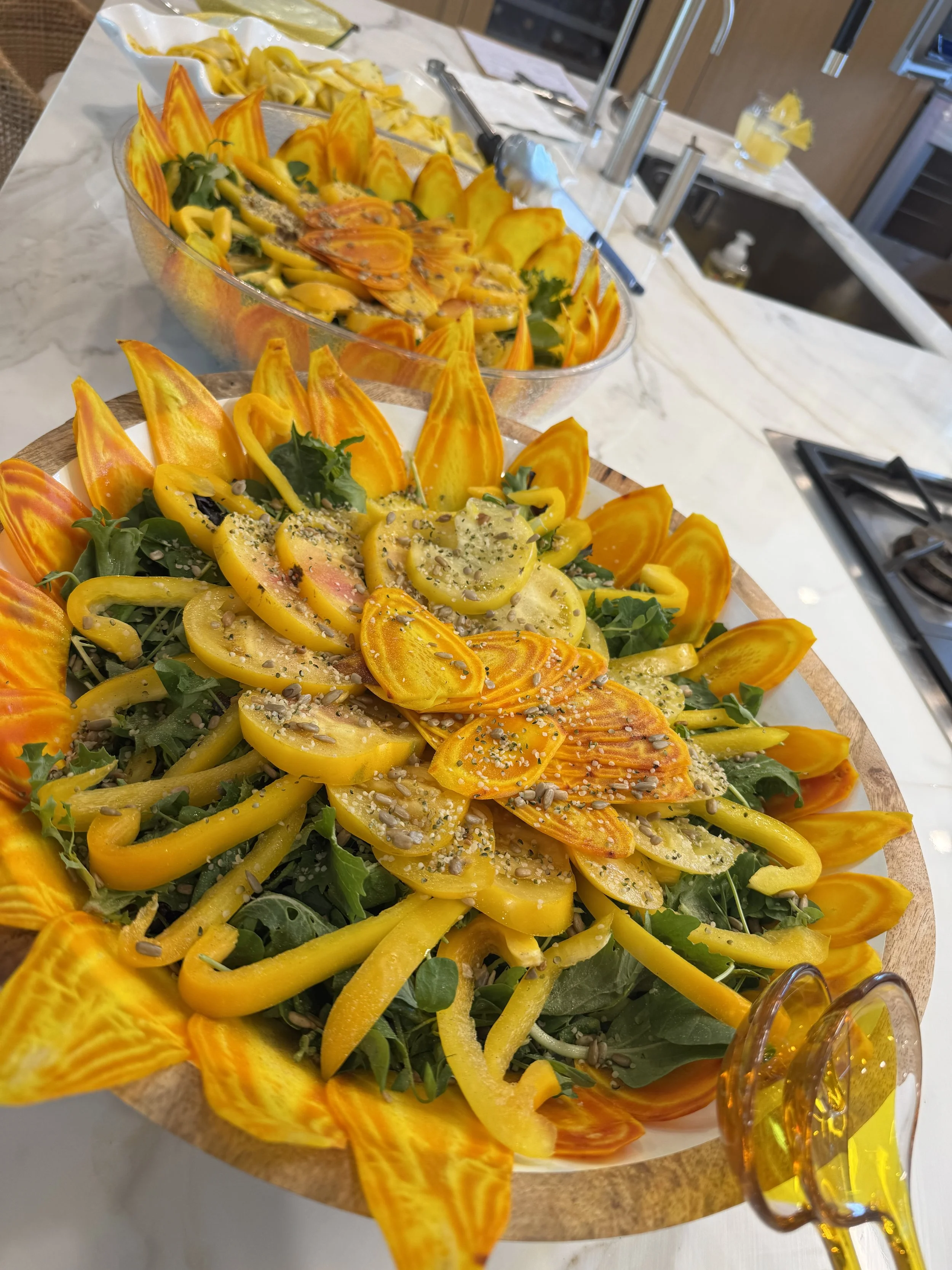 Colorful salad with yellow and orange bell peppers, greens, sunflower seeds, and a variety of sliced vegetables on a wooden platter, served on a marble countertop.