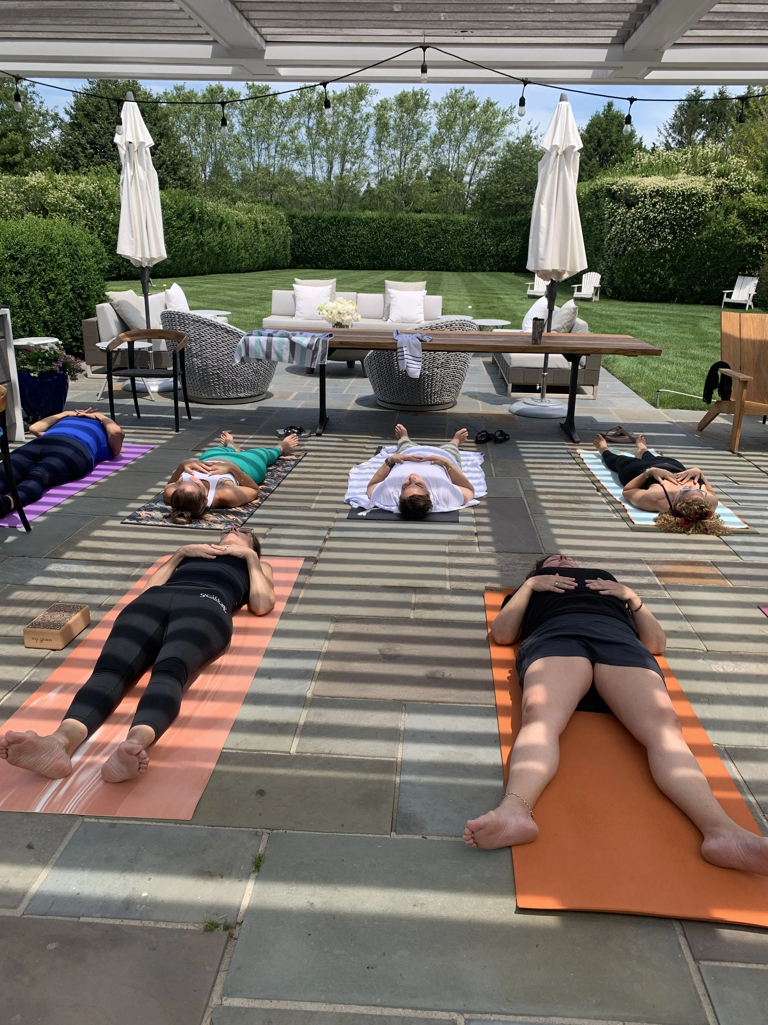 People lying on yoga mats outdoors under a covered patio, practicing meditation or relaxation.