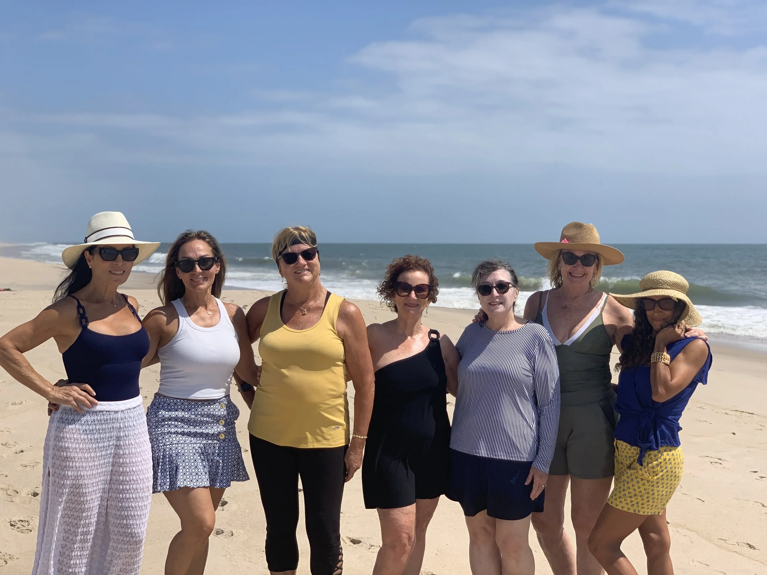 Group of eight women standing on a sandy beach near the ocean, wearing summer clothes and wide-brimmed hats, smiling, with blue sky and clouds in the background.