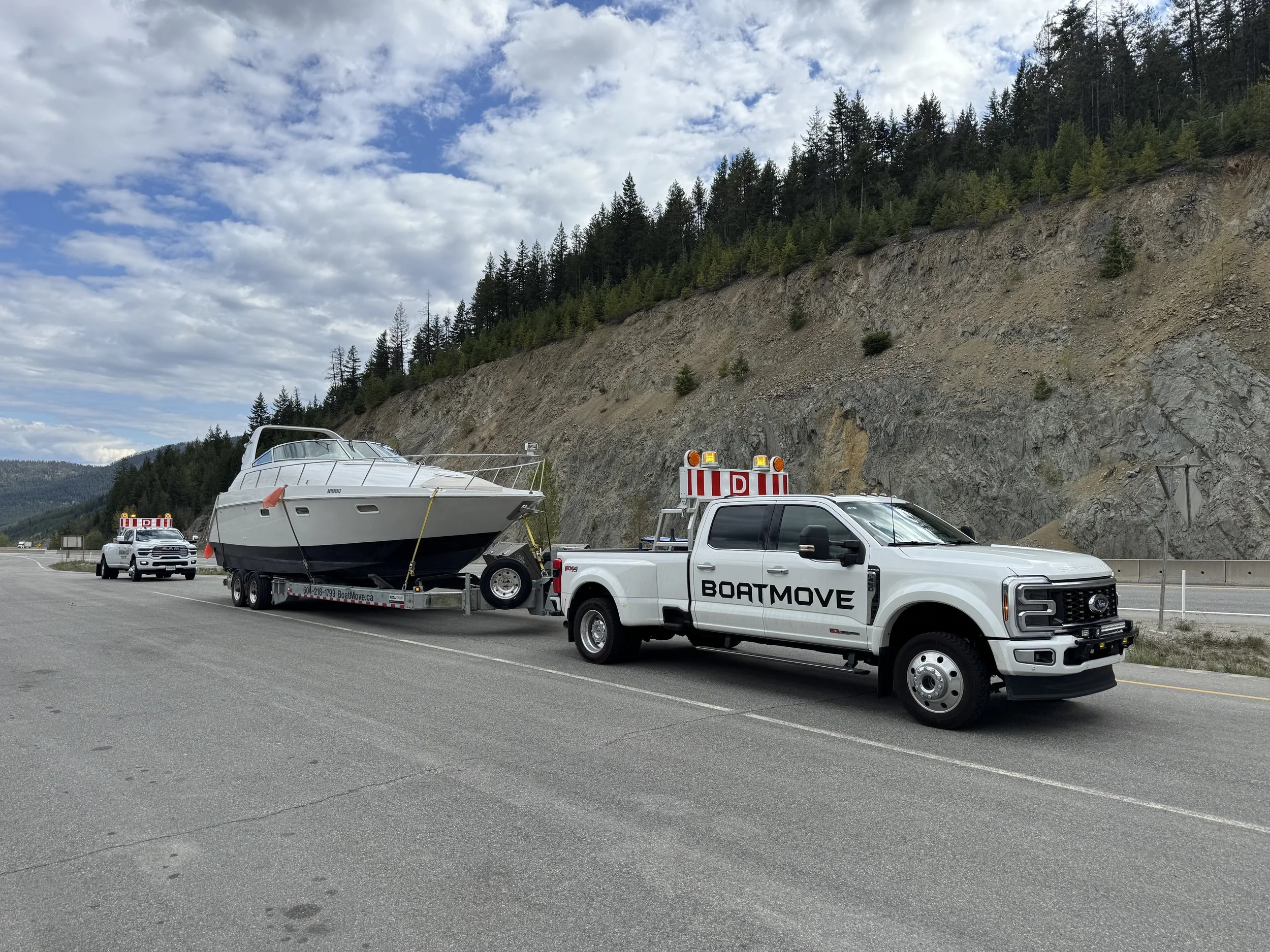 A boat on a trailer being towed by a white pickup truck on a highway, with a backdrop of forested hills and a partly cloudy sky.