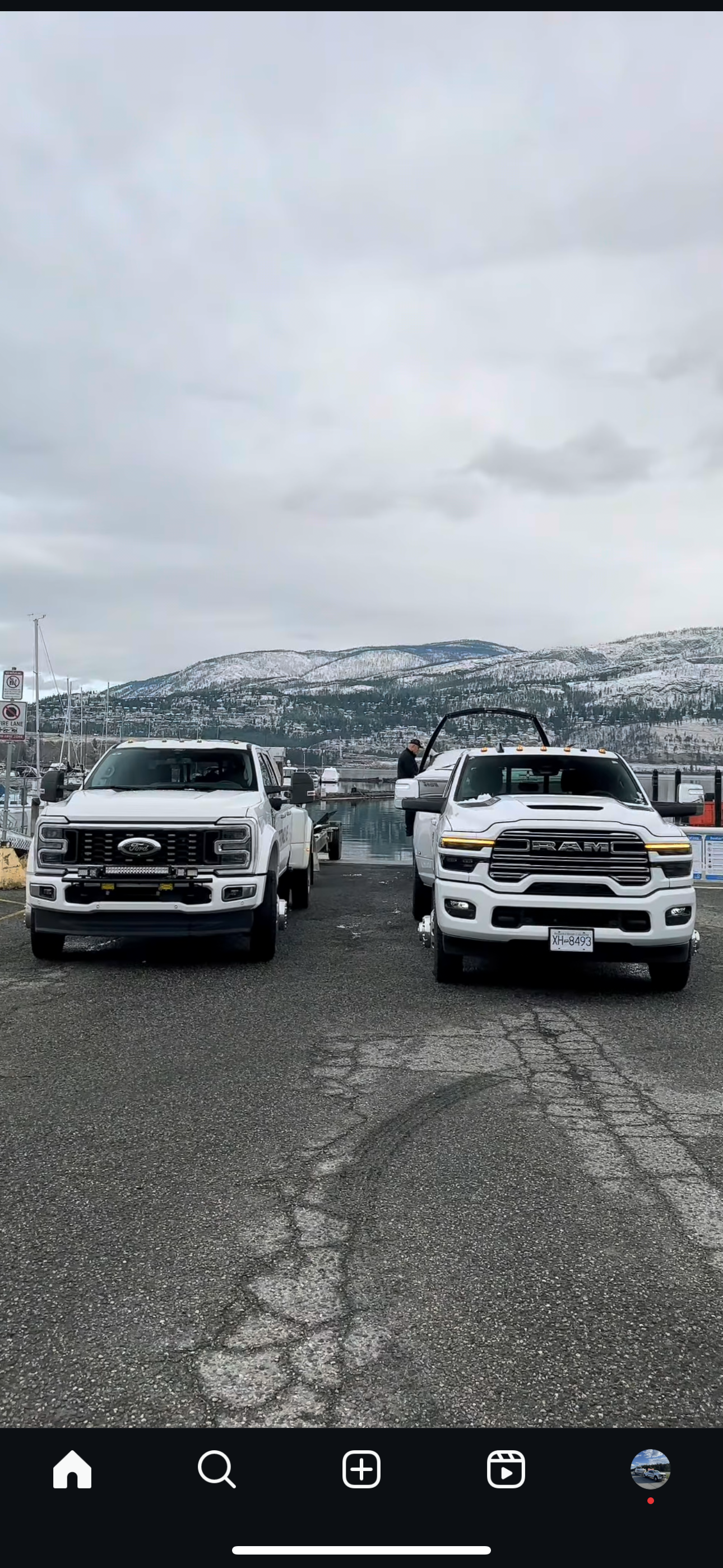 Two white trucks parked on a paved lot with a body of water and snow-covered mountains in the background, overcast sky.