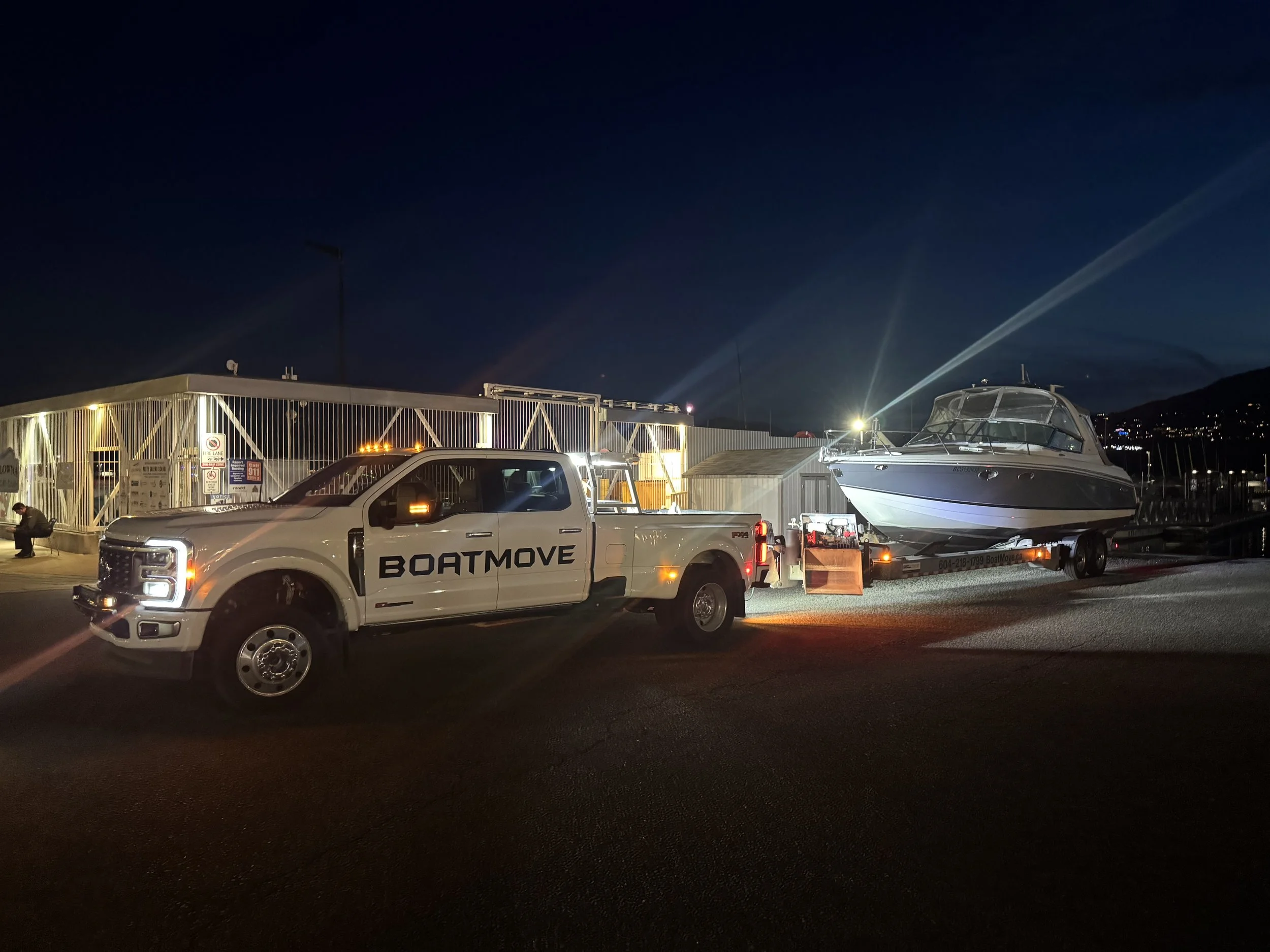 A nighttime scene at a marina with a boat on a trailer, a pickup truck marked with 'BOATMOVE', and a hold with bright lights and safety signs.