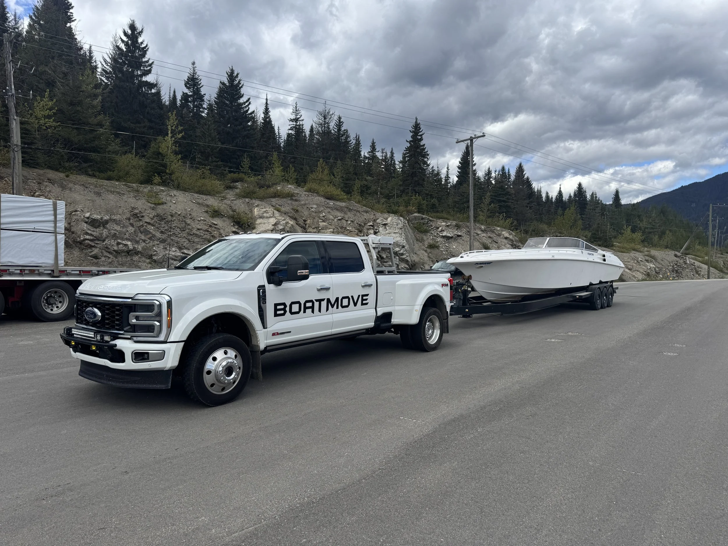 A white pickup truck towing a boat on a trailer parked on a paved road with hillside and dense evergreen trees in the background under a cloudy sky.