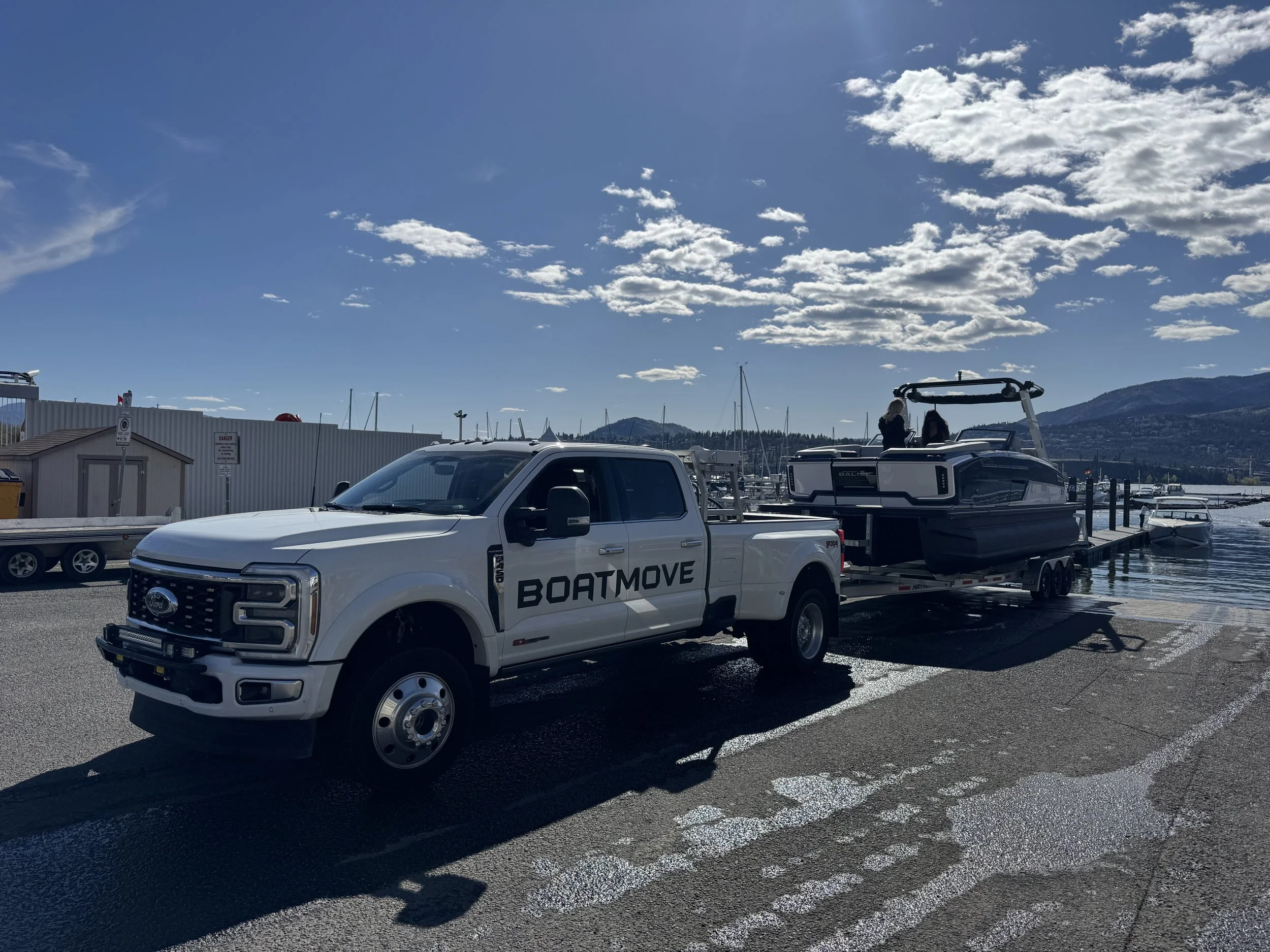 A white Ford pickup truck with a boat trailer attached, labeled 'BOATMOVE', parked by the water under a partly cloudy sky, with yachts and mountains in the background.
