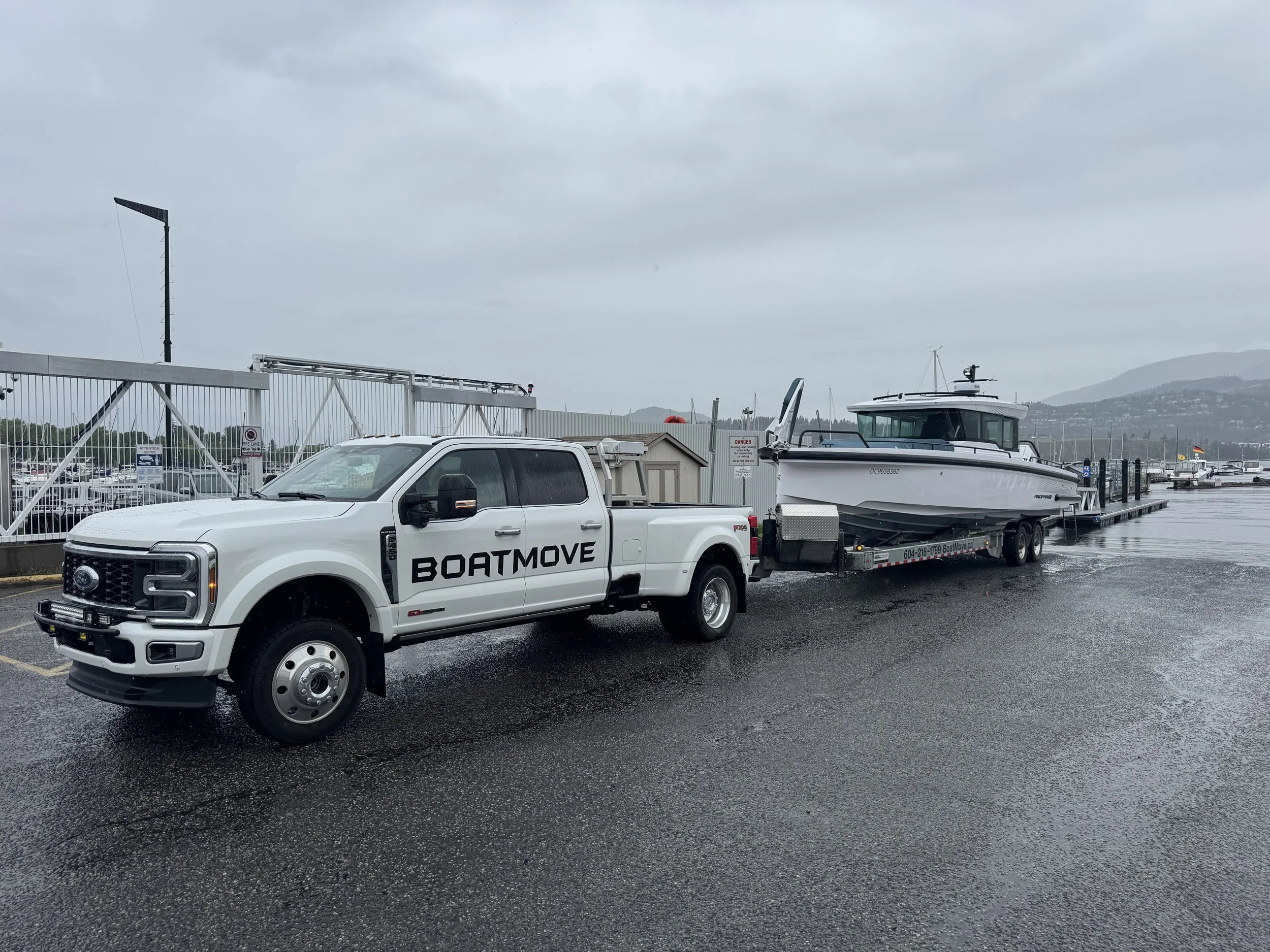 A white pickup truck with the word 'BOATMOVE' printed on the side is parked next to a boat on a trailer at a dock or marina. The scene is during a cloudy day with overcast skies, and there are hills in the background.