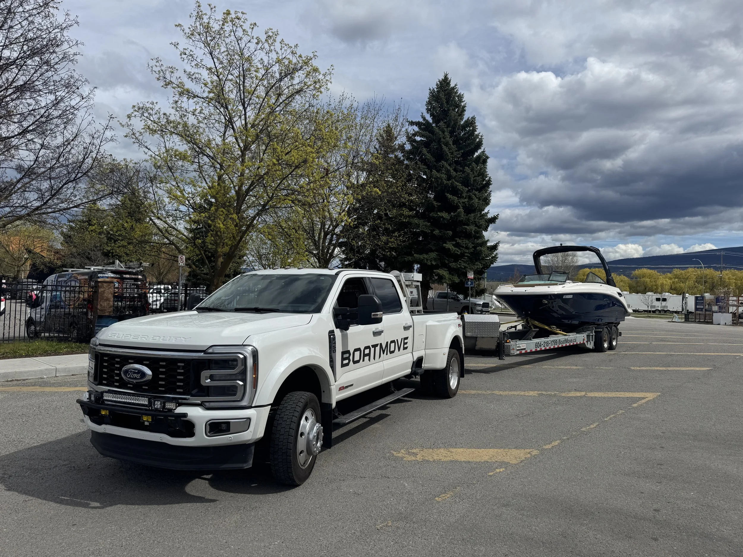 A white Ford Super Duty pickup truck with a BoatMove logo on the side, attached to a boat trailer carrying a boat, parked in a parking lot with trees and dark clouds in the sky.