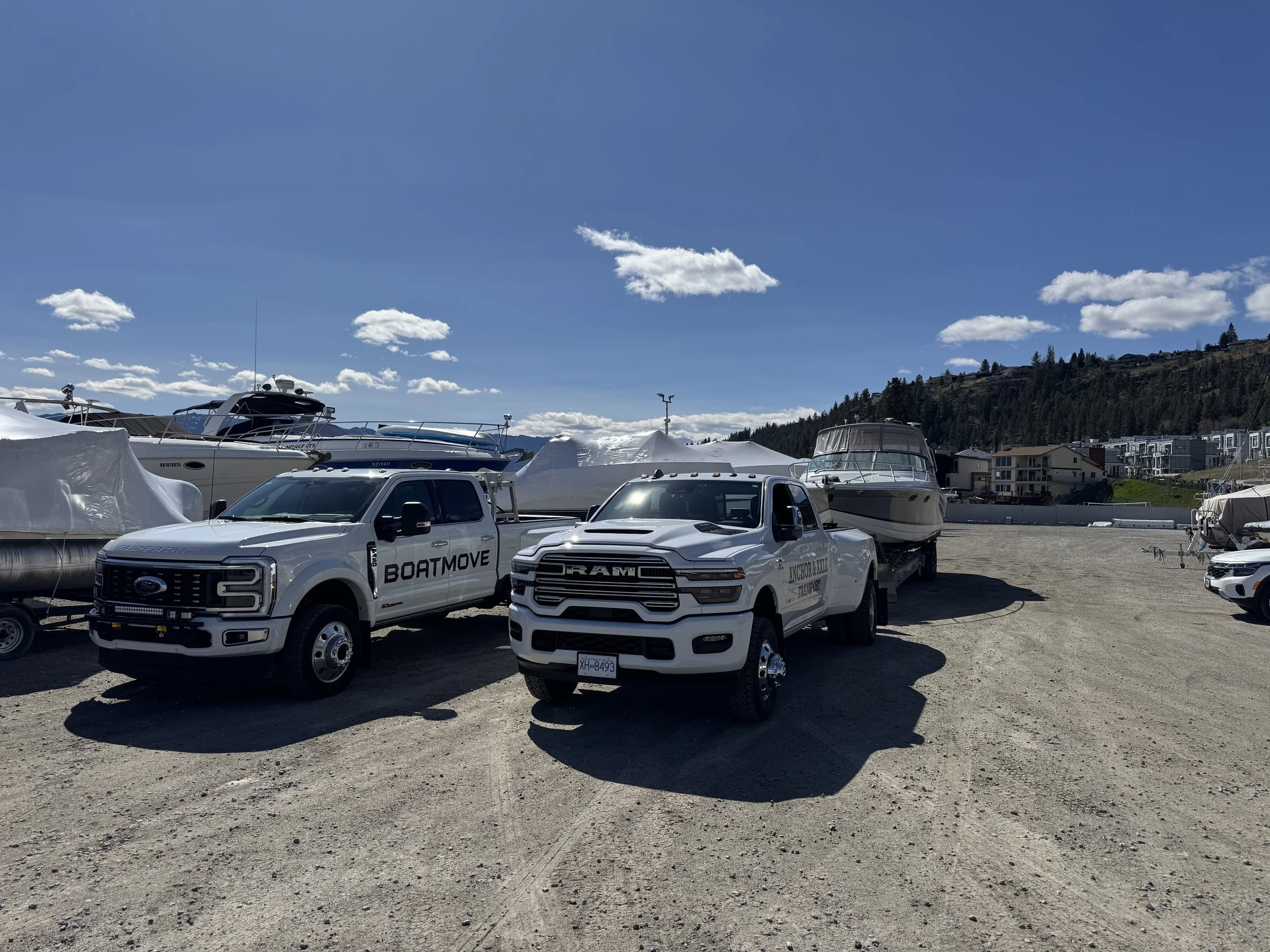 A marina with several boats docked, some covered with white tarps, and two white trucks in front with logos 'BoatMove' and 'Anchor & Lake Transport,' under a blue sky with clouds and a hillside with buildings and trees in the background.